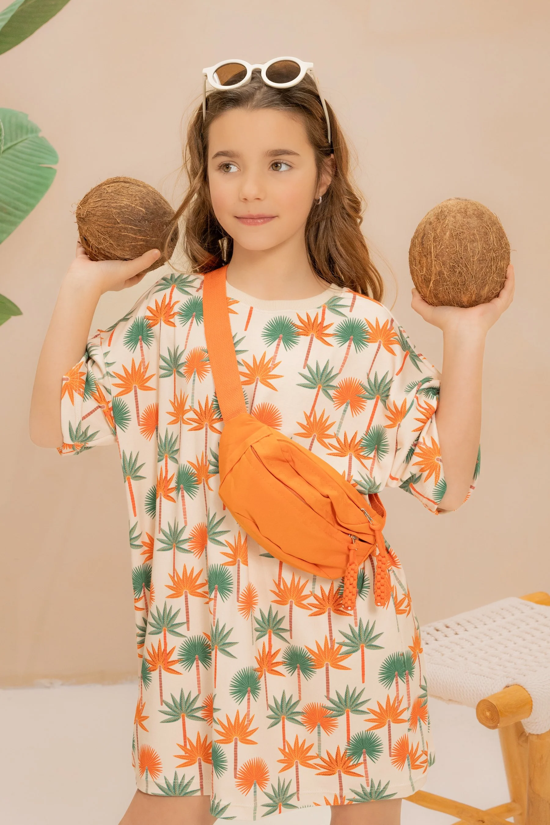 Une jeune fille porte une robe colorée avec un motif de palmiers vert, orange et blanc, ainsi qu'un sac banane orange, elle tient deux noix de coco, une dans chaque main, et porte des lunettes de soleil blanches sur la tête.