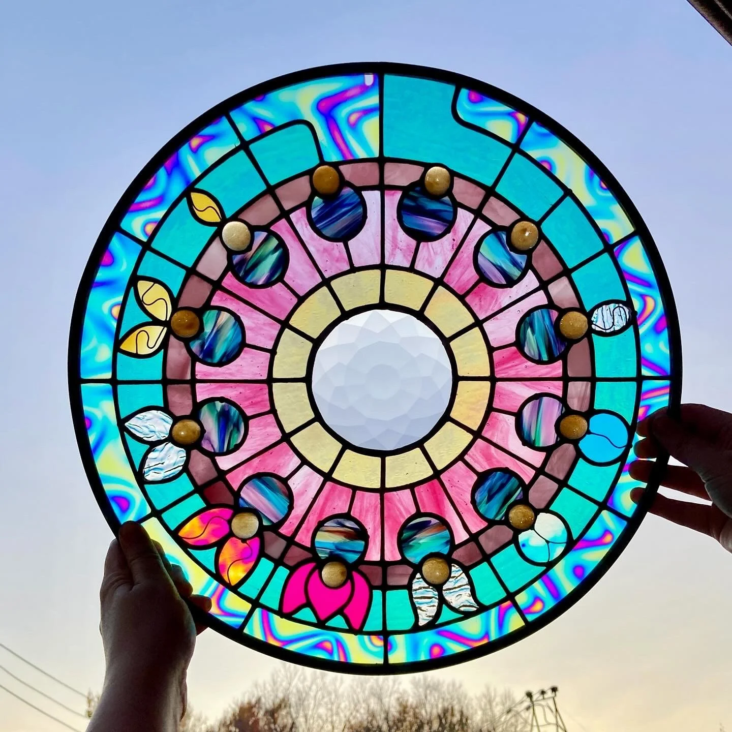 Vitrail coloré avec des motifs floraux, tenu par deux mains contre un ciel clair.