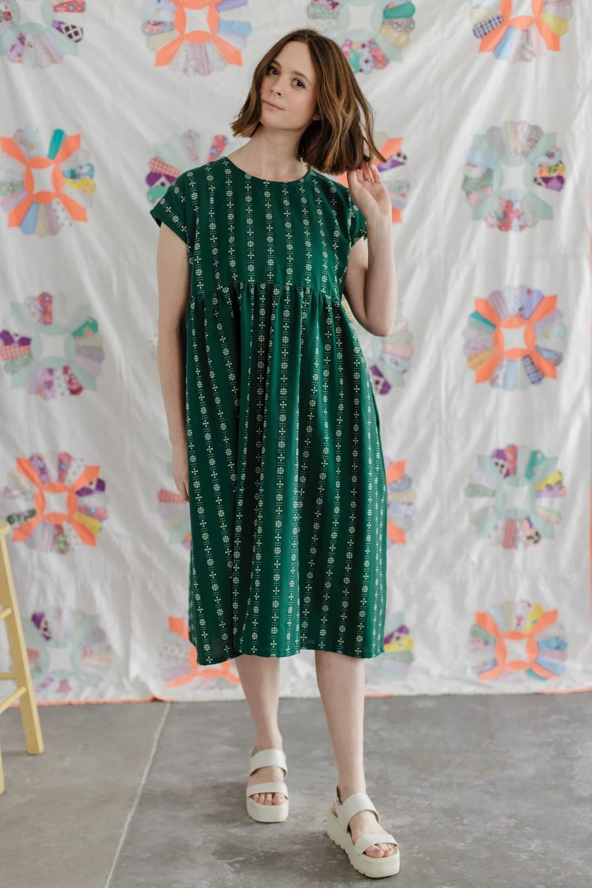 Une femme porte une robe verte à motifs blancs et longs cheveux bruns. Elle se tient devant un drap blanc décoré de motifs colorés en forme d'étoile. La femme porte des sandales blanches à plateforme.