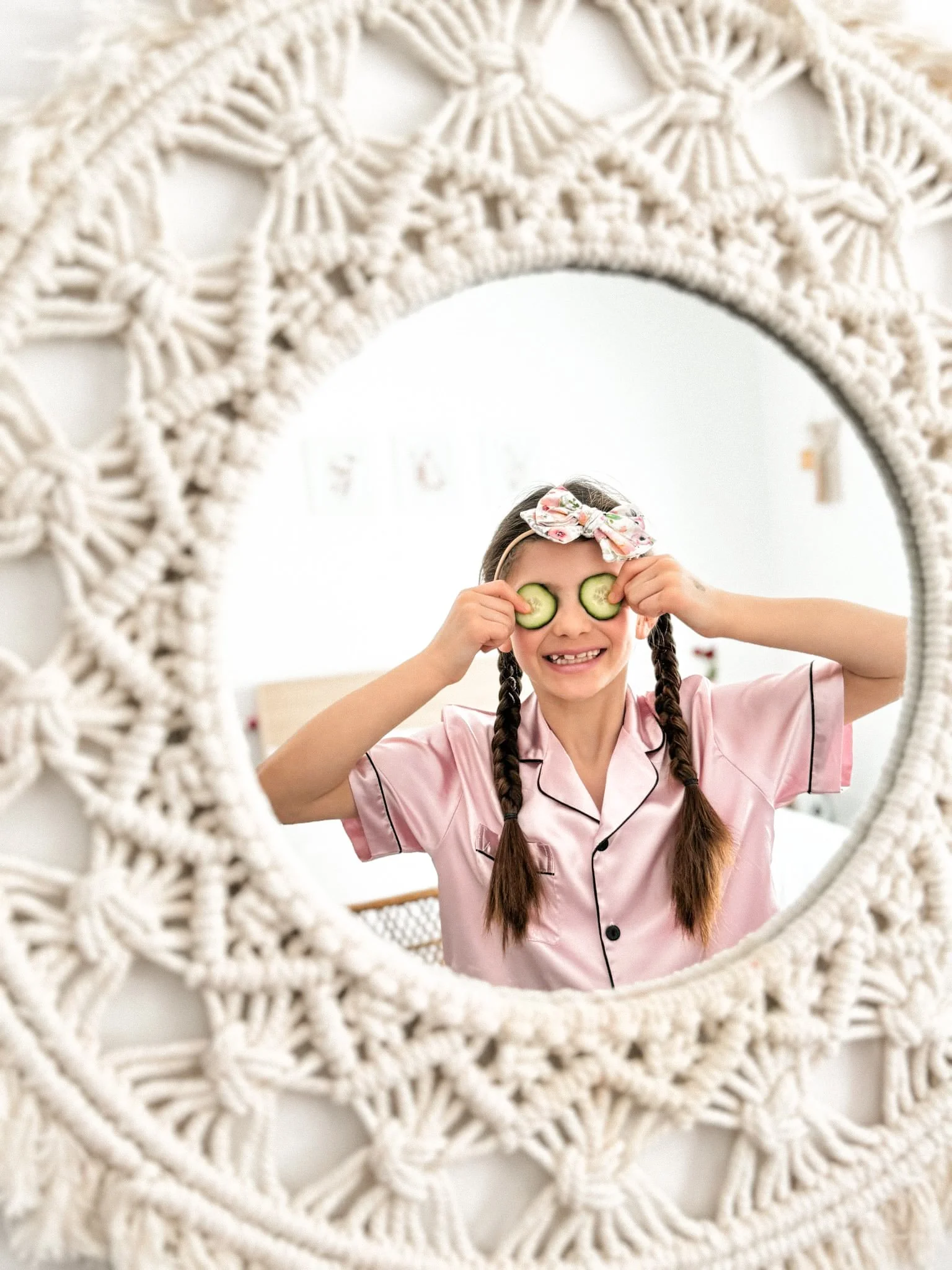 Jeune fille posant devant un miroir avec un cadre en macramé blanc, portant un peignoir rose et des lunettes de concombre, souriant.