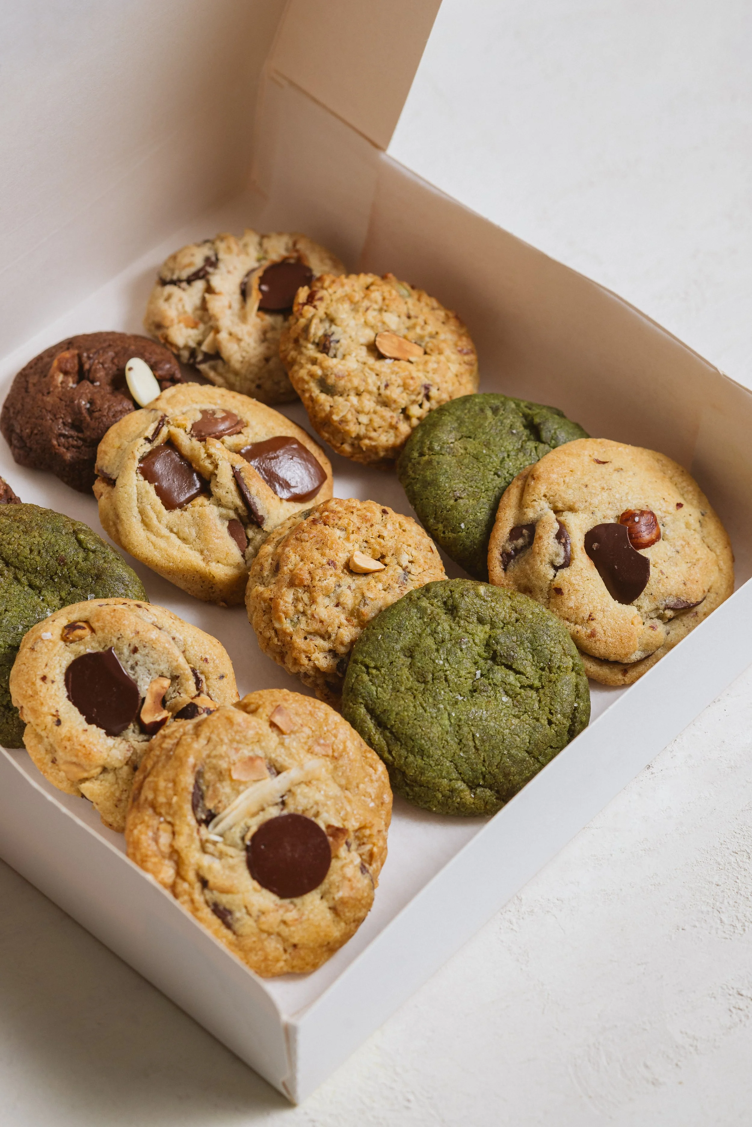 Gamme de cookies aux pépites de chocolat, avec différentes saveurs comme Matcha vert, et autres avec des noix, dans une boîte blanche.