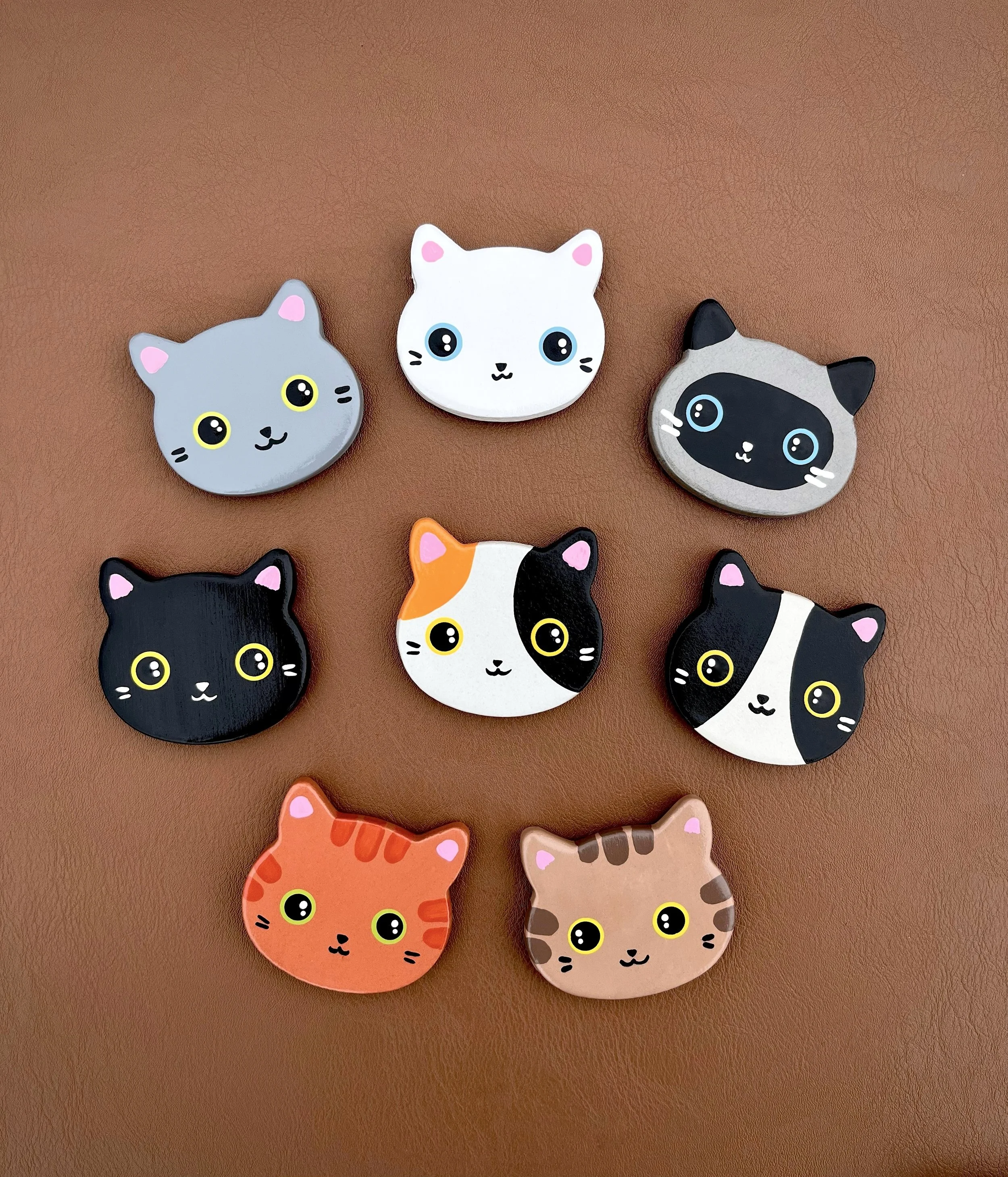 Collection de badges en forme de visages de chats, chacun avec différentes couleurs et motifs, disposés sur une surface marron.