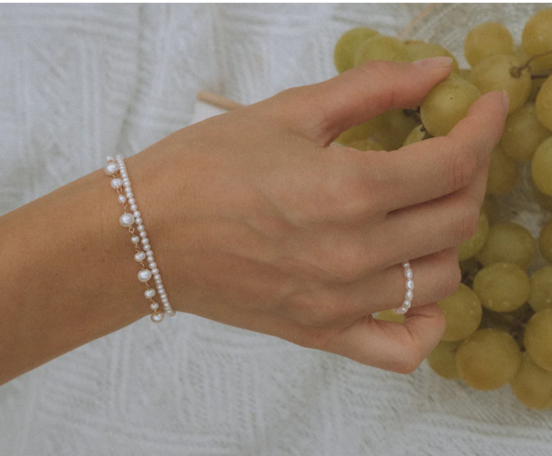 Une main portant un bracelet et une bague, tenant un raisin vert au-dessus d'un panier de raisins verts.