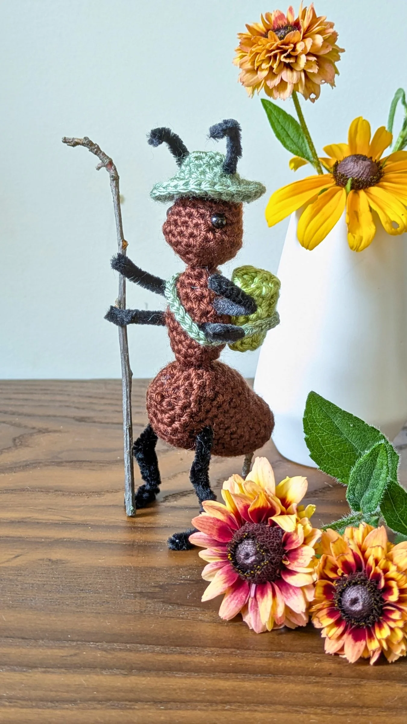 Figure de laine humanoïde avec un chapeau, un bâton, et un sac, entourée de fleurs de tournesol dans un vase et sur une table en bois.