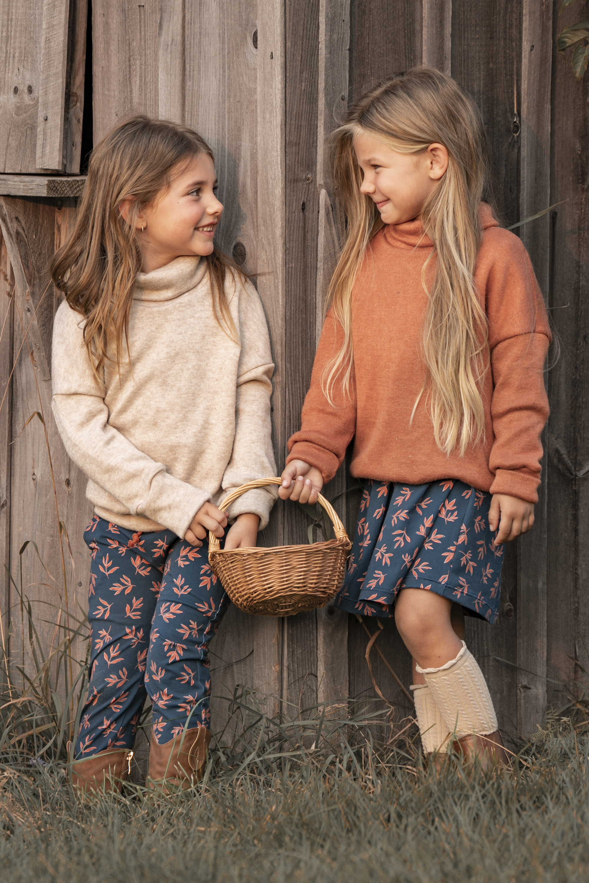 Deux jeunes filles souriantes, tenant un panier, debout devant une clôture en bois, portant des vêtements d'automne avec des motifs floraux.
