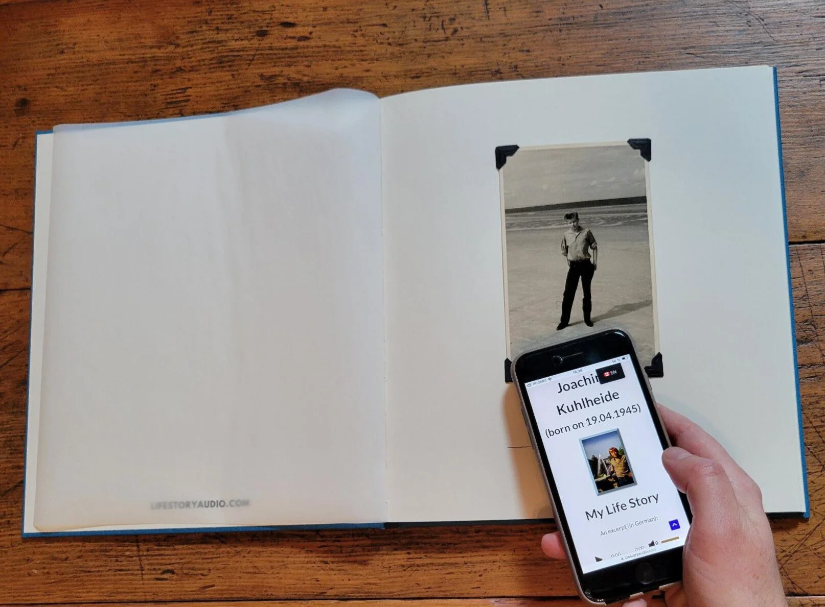 Une personne tient un téléphone avec une application montrant la biographie de Joachim Kulheide devant une photo en noir et blanc d'un homme sur une plage. La page est ouverte dans un livre à côté d'une table en bois.