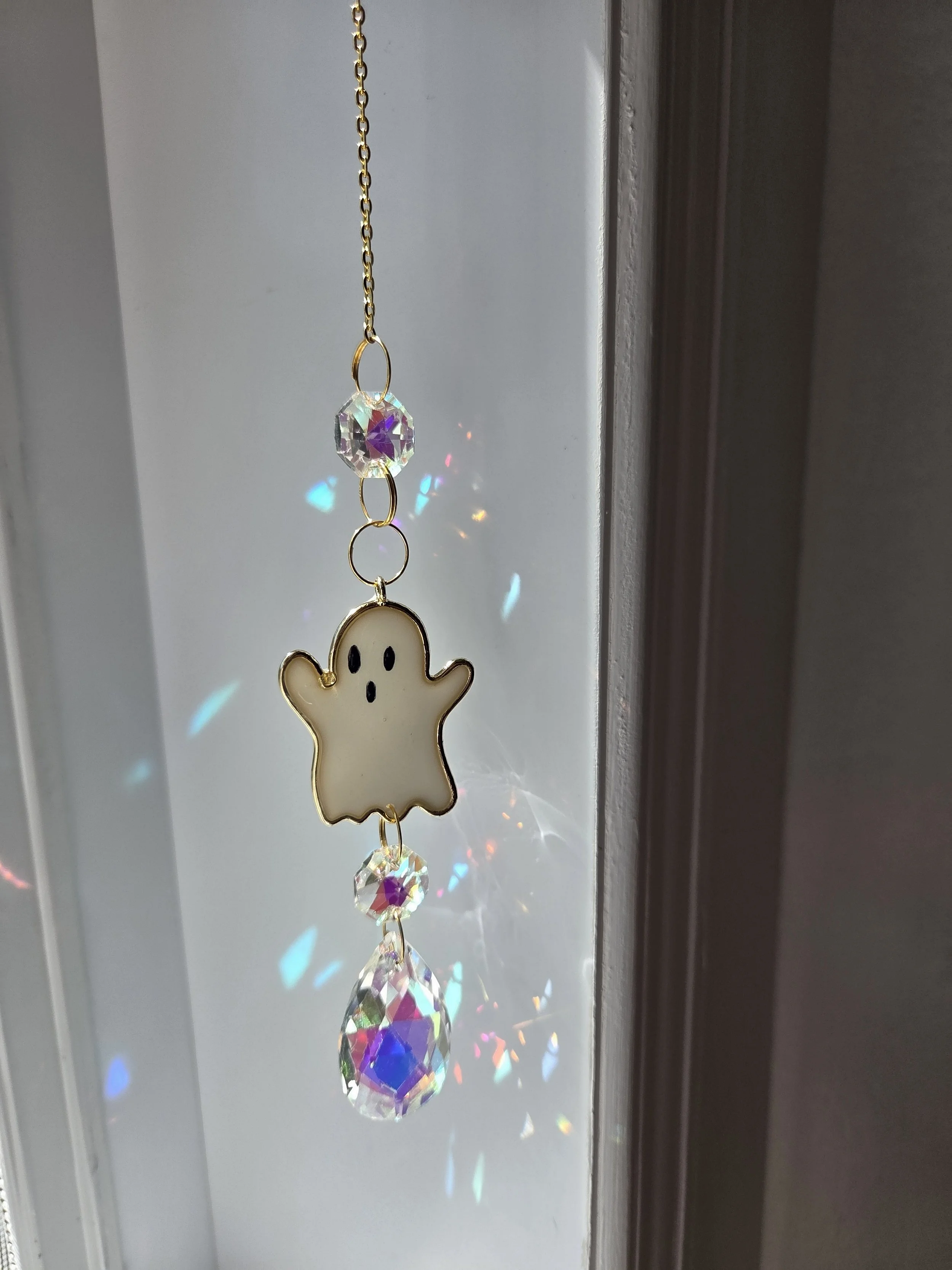Décoration suspendue avec un pendentif en forme de fantôme, en plastique blanc avec des yeux noirs, accompagné de cristaux colorés et transparents qui créent des reflets de lumière.