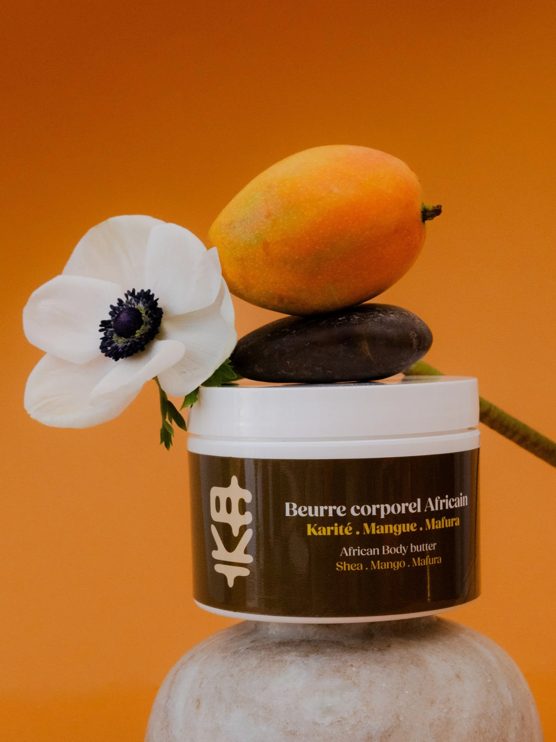 Une boîte de beurre corporel africain avec un fond orange, une pierre et une fleur blanche.