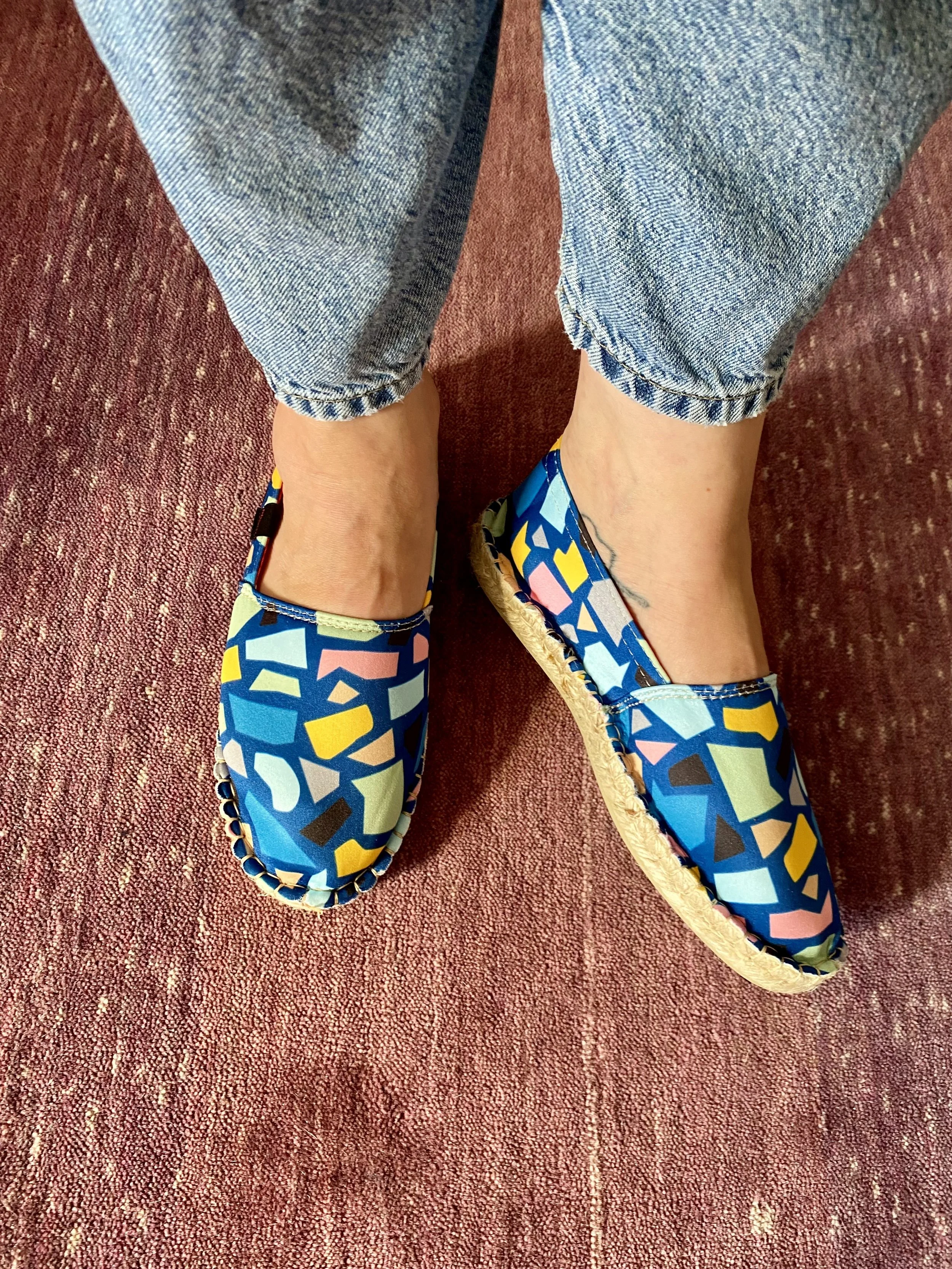 Personne portant des chaussures colorées avec un motif géométrique bleu, jaune, rose, vert et noir, avec un espace en caoutchouc naturel, en jean bleu avec un ourlet roulé, assise sur un tapis rouge.