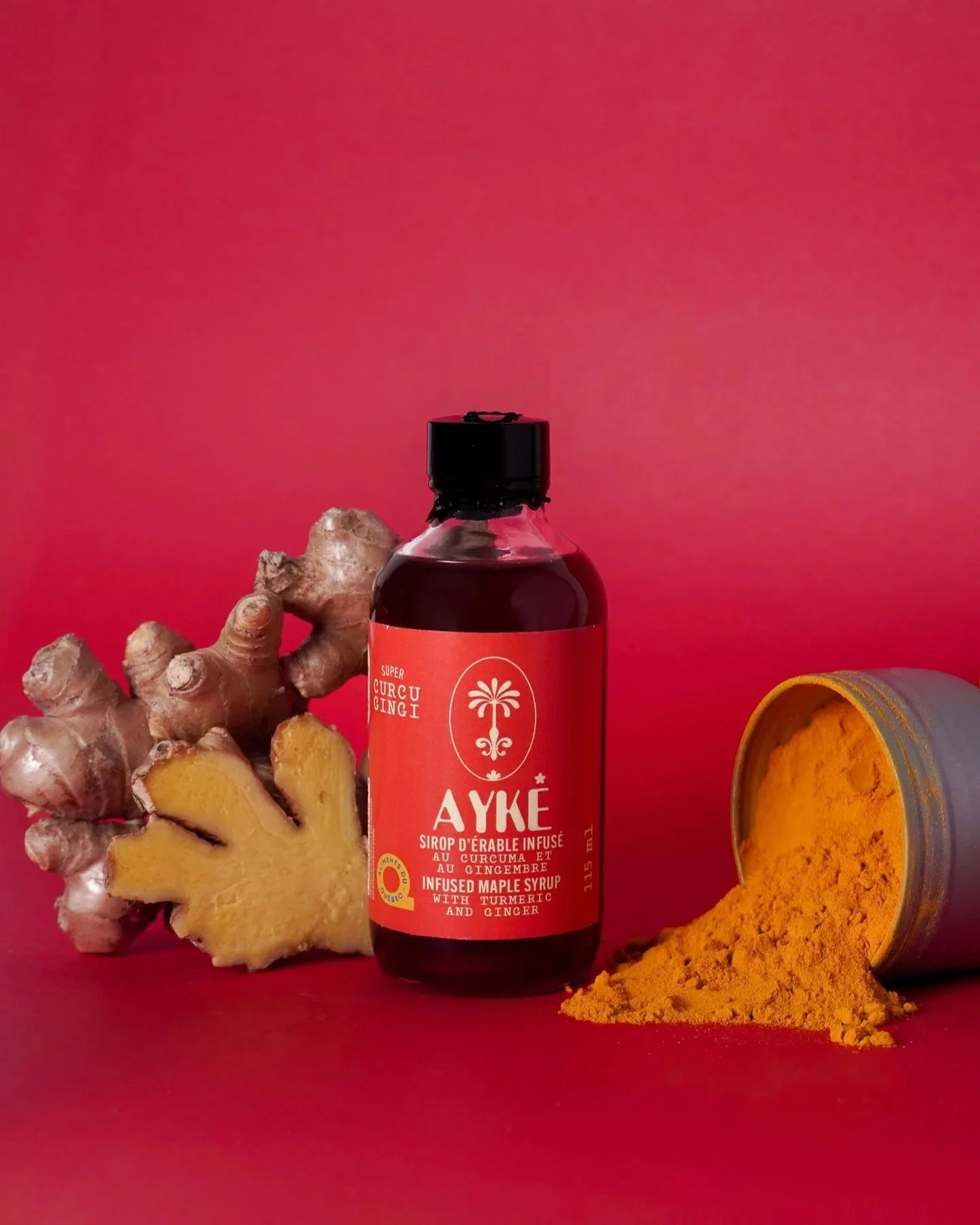 Une bouteille de sirop d'érable infusé au curcuma et au gingembre, un morceau de gingembre frais, du curcuma en poudre dans un pot, et un fond rouge.