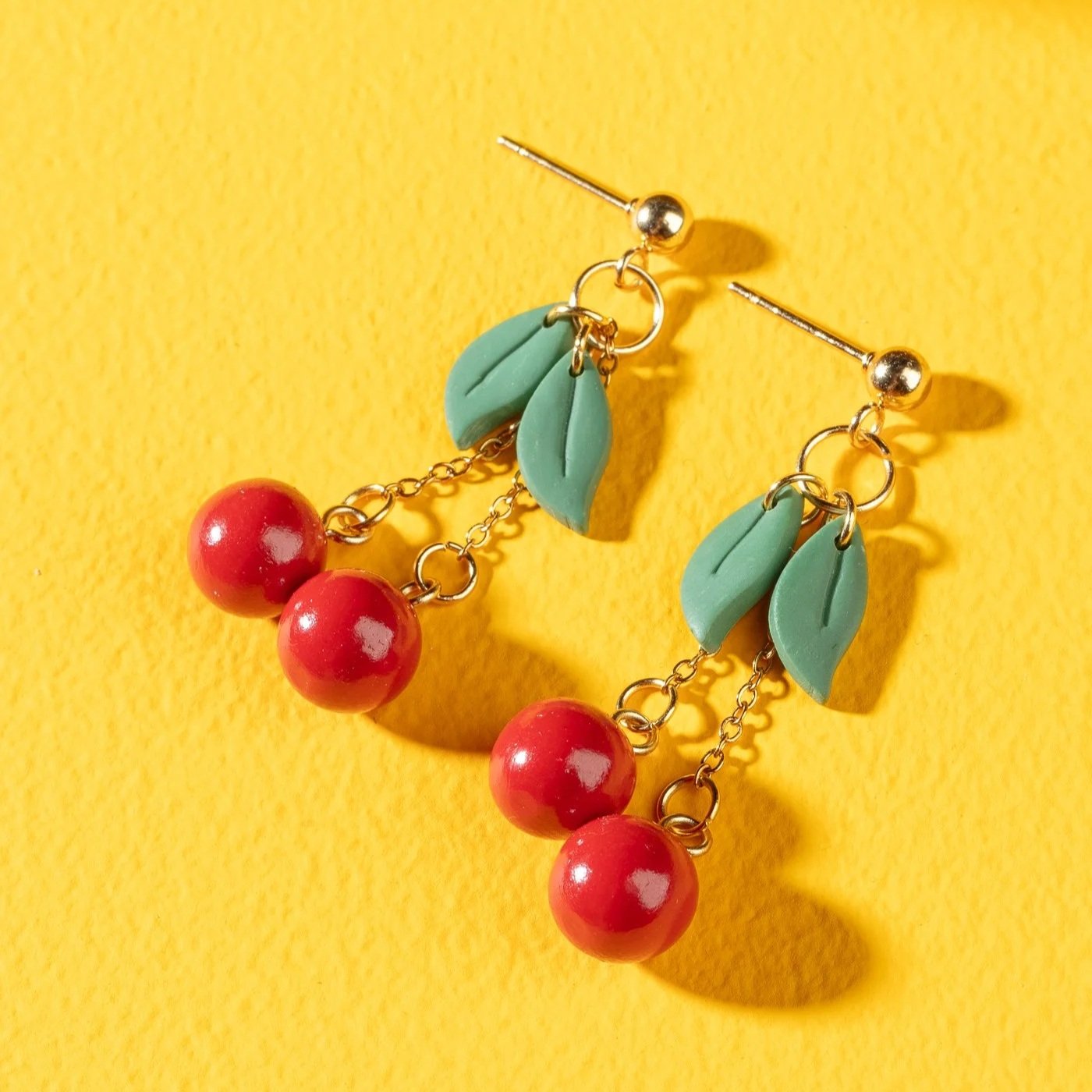 Boucles d'oreilles colorées avec des perles rouges, des feuilles en plastique vert et des éléments en métal doré, posées sur un fond jaune.