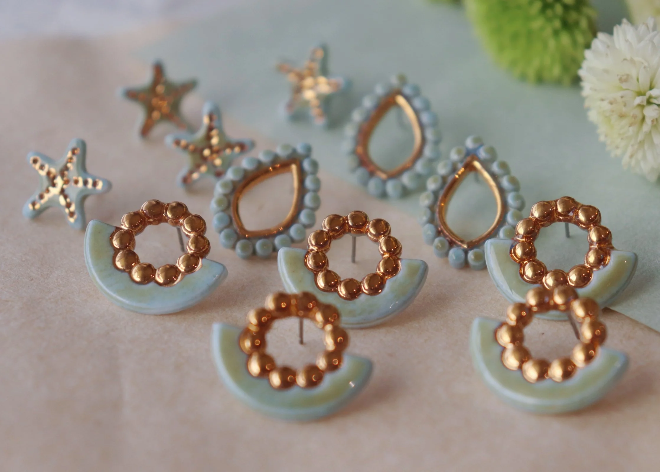 Boucles d'oreilles en forme de croissant et d'étoile, décorées de perles de couleur or et blanche, posées sur une surface neutre avec une fleur blanche en arrière-plan.