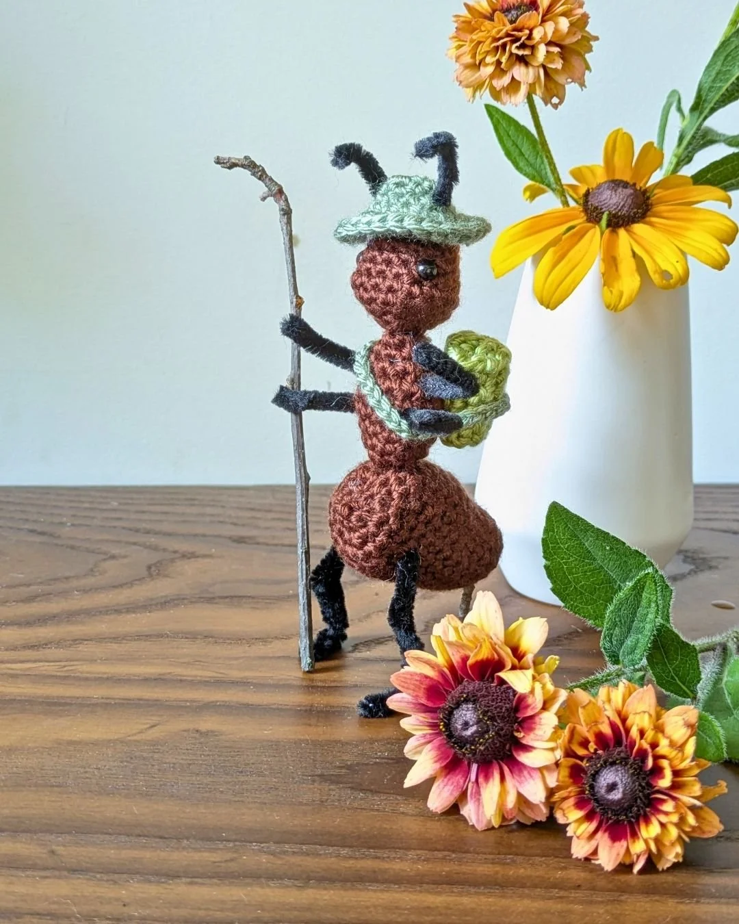 Une figurine de fourmi en crochet portant un chapeau avec des cornes, tenant une baguette, située sur une table en bois avec un vase blanc contenant des fleurs jaunes et oranges.