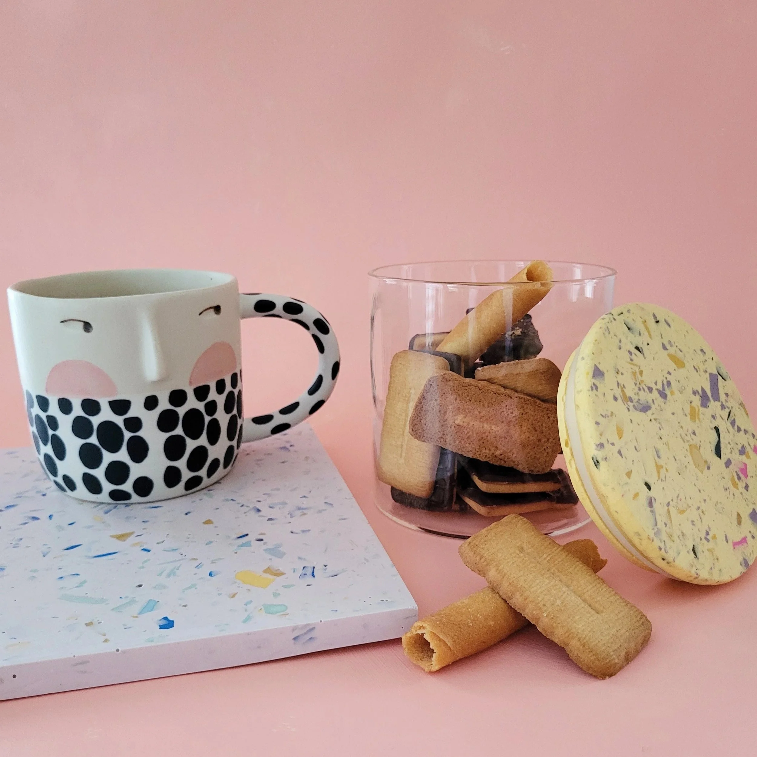 Une tasse, un verre avec des biscuits, et des macarons, sur une table rose, contre un mur rose.