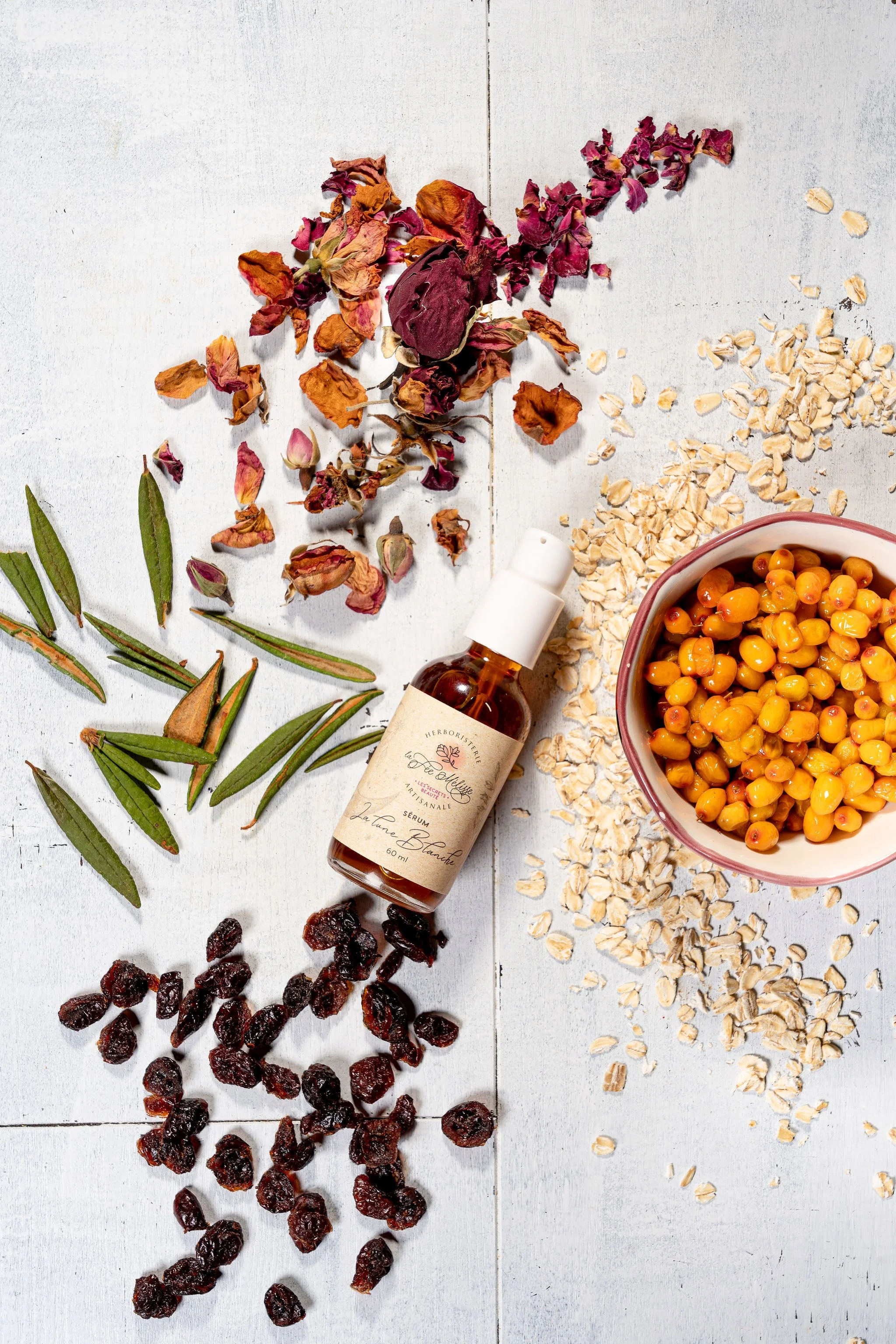 Présentation d'ingrédients pour une tisane ou infusion : pétales de rose séchés, feuilles de plante aromatique, fruits séchés, flocons d'avoine, barbe à papa, bouteilles de sérum, fourrure de fruit ou baie séchée, et un bol rempli de baies d'aron.