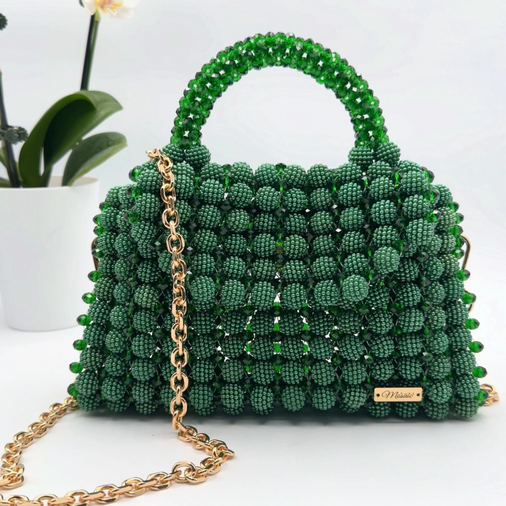 Un sac à main vert composé de petites perles en forme de fleurs, avec une chaîne en or rosé, posé à côté d'une plante dans un pot blanc.
