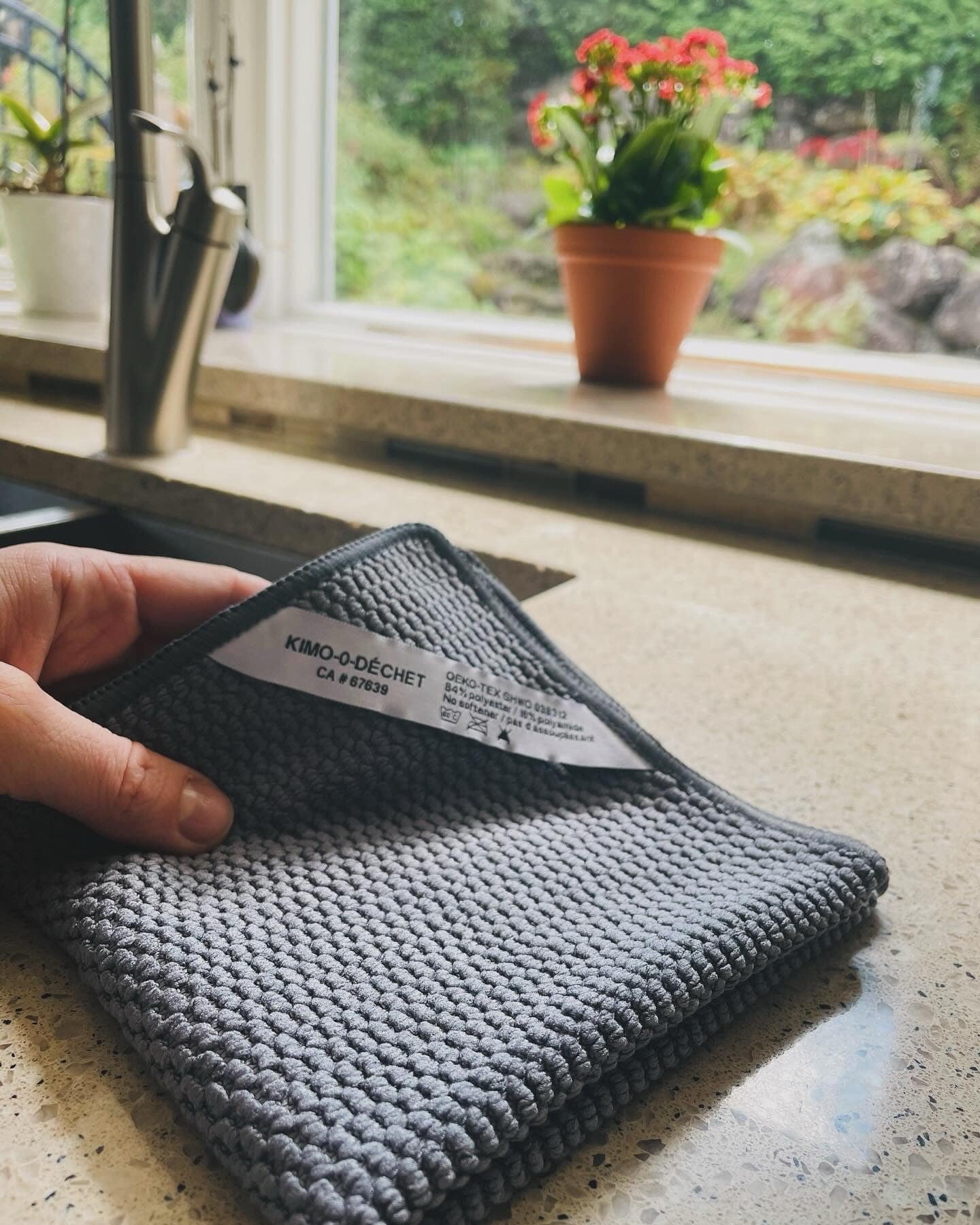 Une main tient un porte-cartes en tissu gris sur une table en quartz. À l'arrière-plan, une fenêtre avec vue sur un jardin avec des plantes et des rochers.