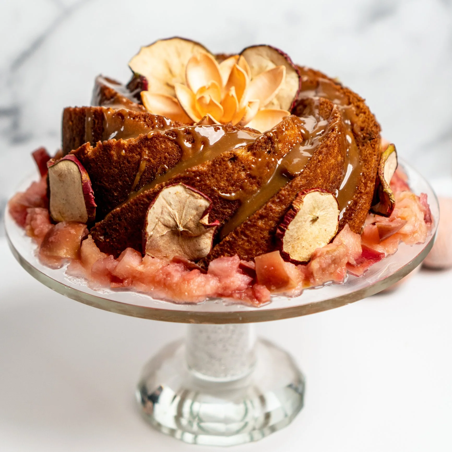Gâteau décoré avec des tranches de pomme déshydratée, de la pâte d'amande en forme de fleur et une sauce caramel, présenté dans un verre à pied.