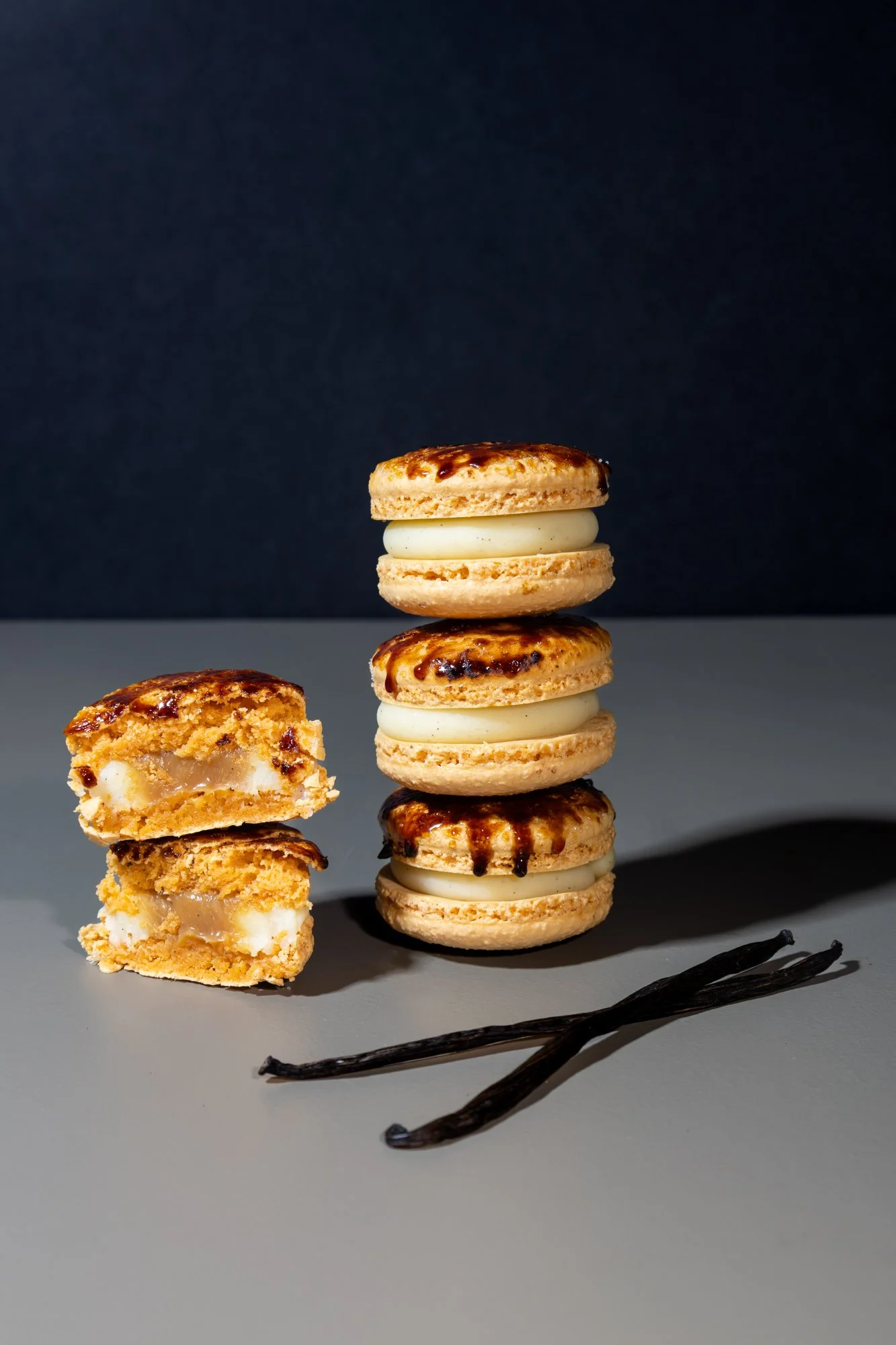 Un tas de macarons avec une moitié cassée, une vanille, deux macarons avec un glaçage au chocolat, et deux bâtons de vanille sur une surface grise.