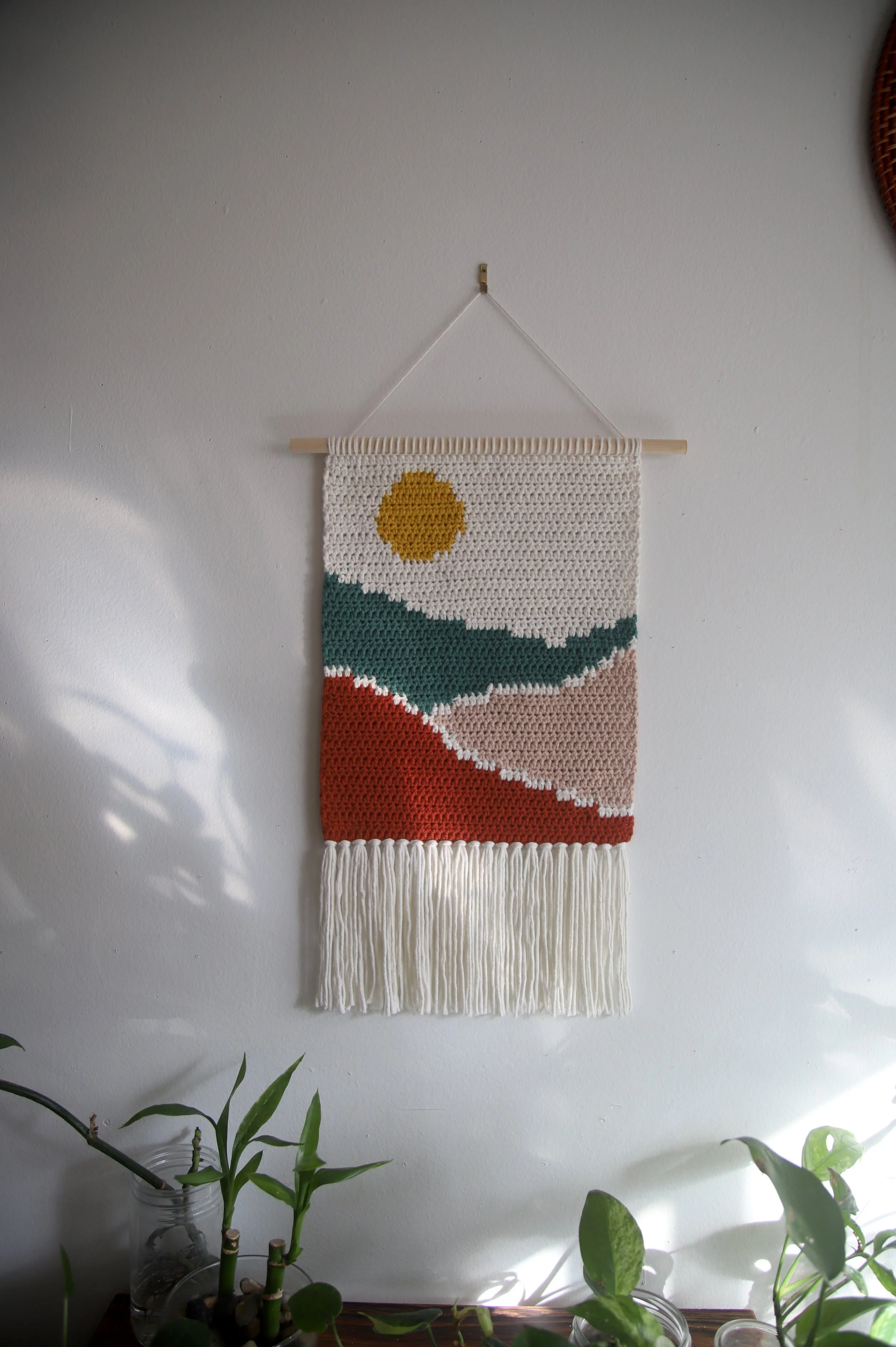 Tapisseries murale colorée avec des motifs de soleil et d'ondulations, suspendue par une corde, décorative, avec des plantes vertes en pot en dessous.