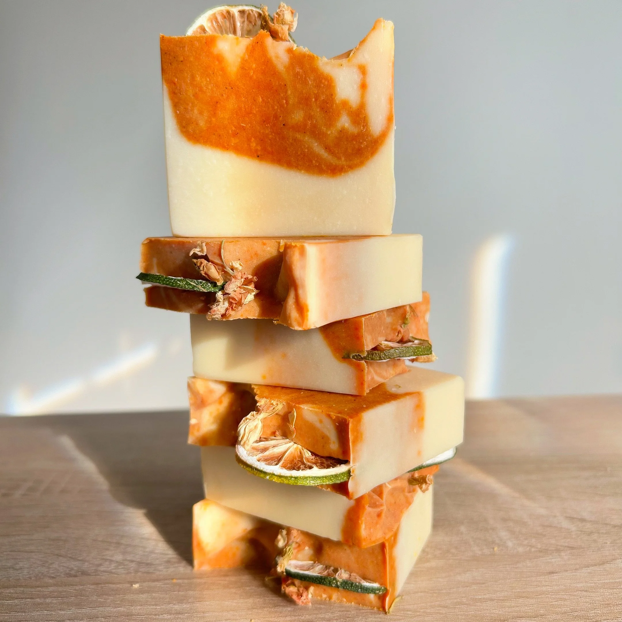 Une tour de plusieurs blocs de savon de différentes couleurs et textures, agrémentée de rondelles de concombre et de fleurs séchées, placée sur une surface en bois, avec un fond neutre.