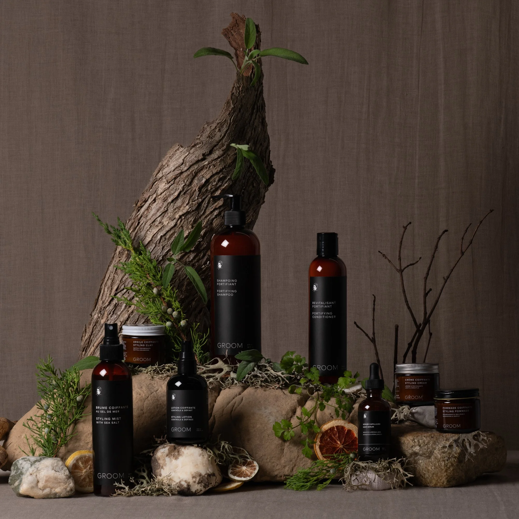 Display de produits de soins capillaires, tels que des shampoings, conditionneurs, et produits coiffants, organisés sur une surface avec des éléments naturels, comme des pierres, des branches, du bois et des plantes, contre un fond en tissu.