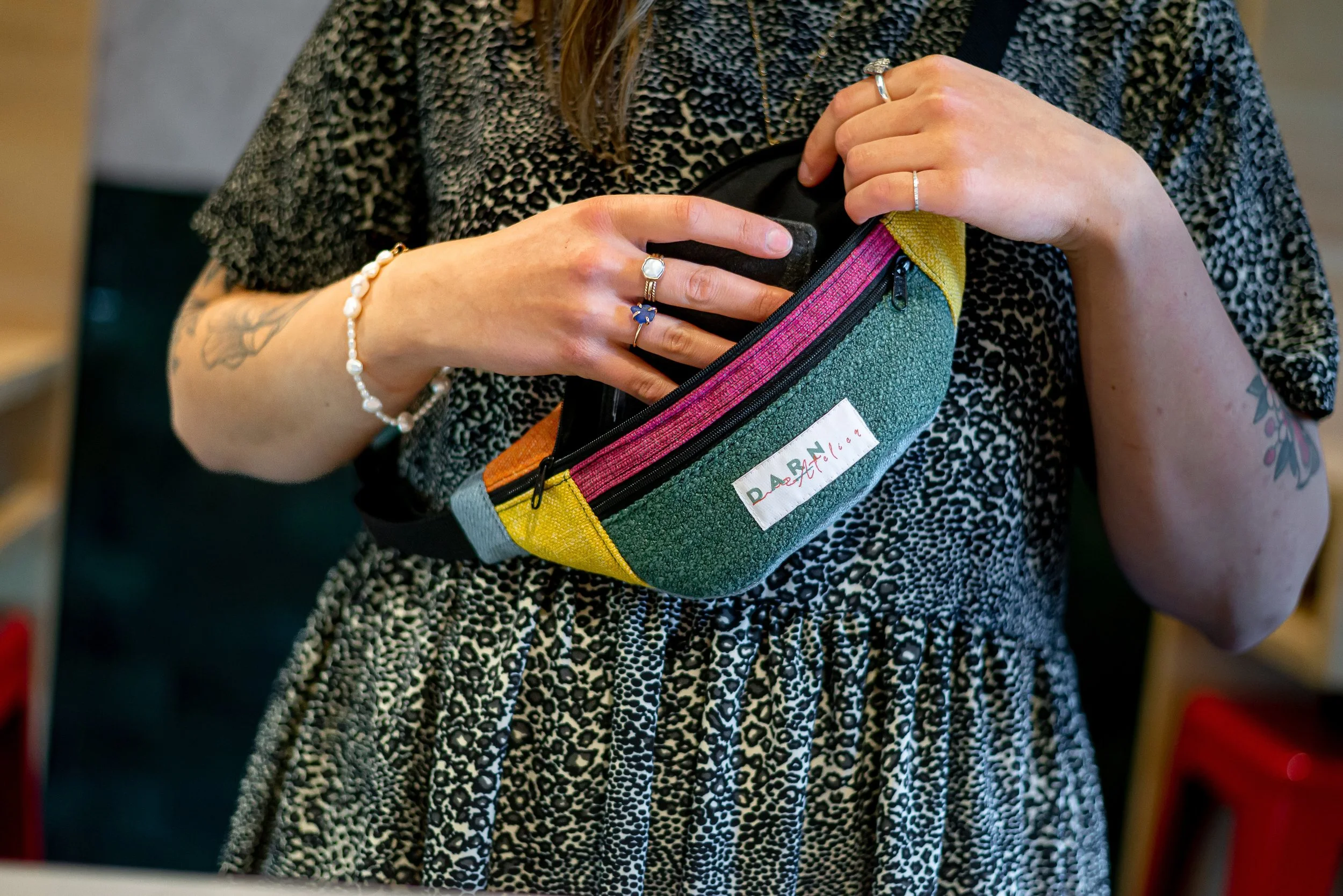 Une personne porte un sac à petite poche colorée vert, rose, jaune et gris, avec une fermeture éclair noire. La personne porte des bijoux dont une bague, une bracelet en perles et une autre bague argentée. La personne porte une robe ou une veste à motif animalier en noir et blanc.