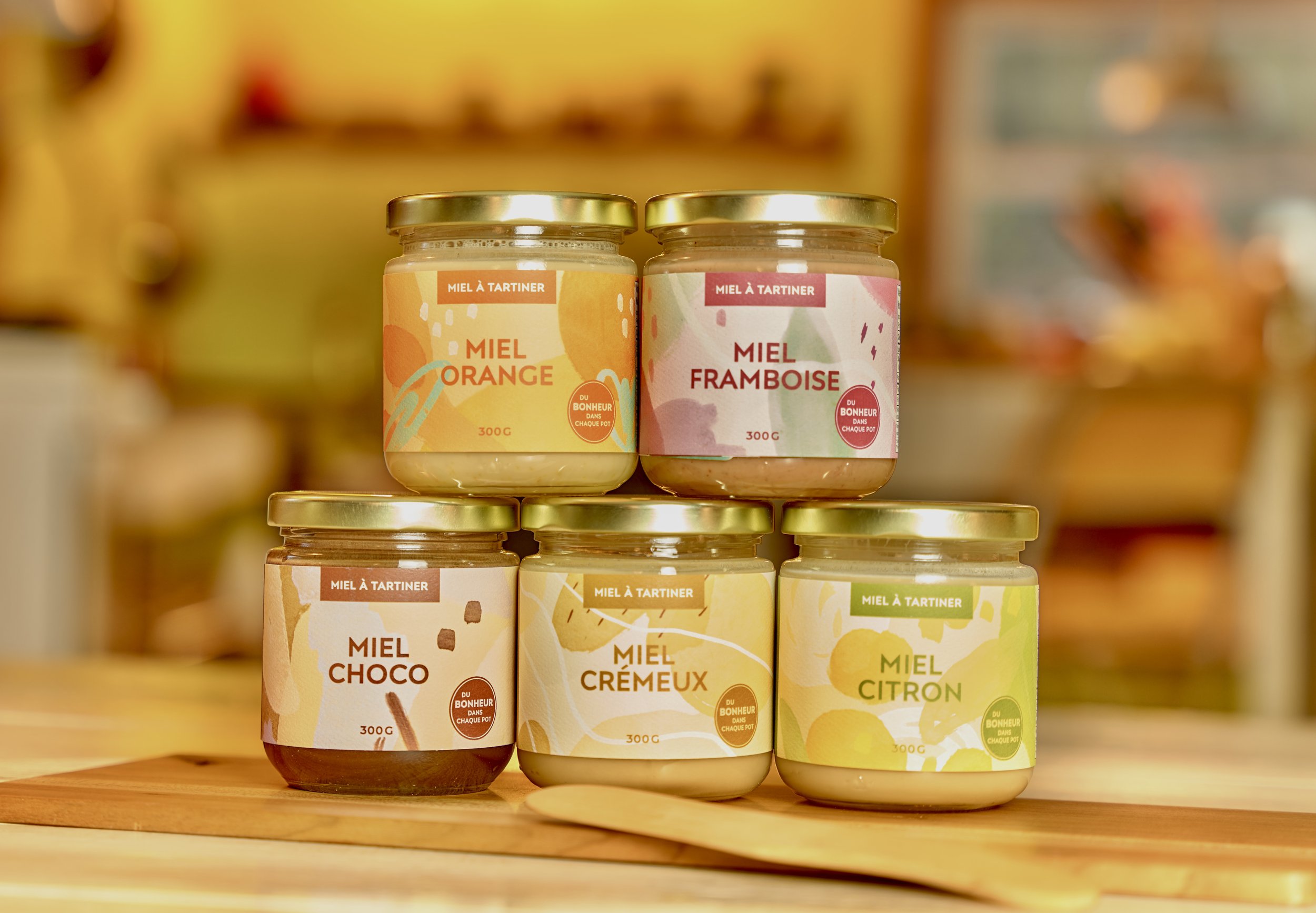Assortiment de pots de miel avec différentes saveurs comme orange, framboise, choco, crèmeux et citron, disposés sur une étagère en bois.