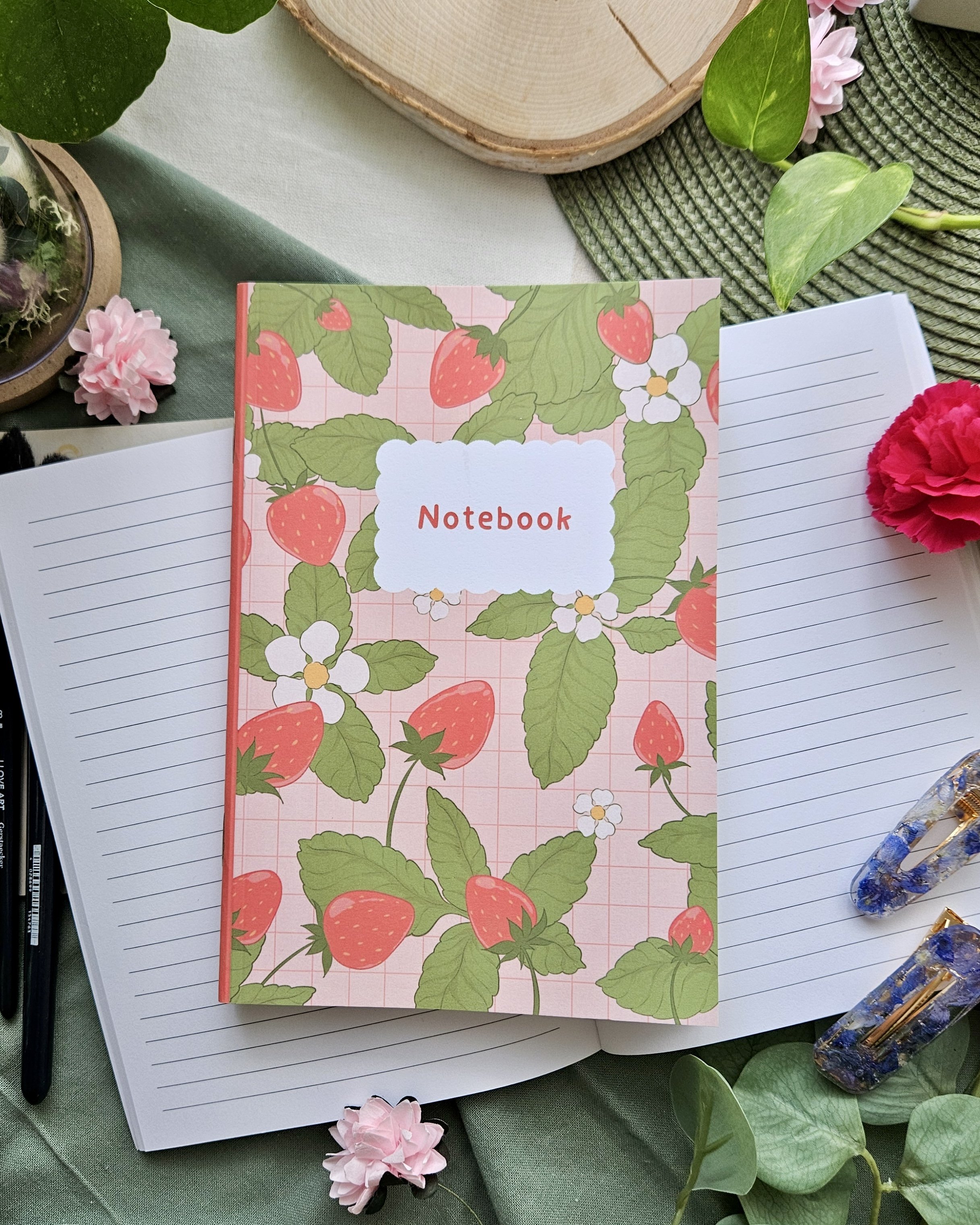 Un carnet vert avec une couverture à motif de fraises, entouré de feuilles vertes, de fleurs en papier roses, d'une plume noire, d'un stylo à bille noir, et de plusieurs pierres décoratives.
