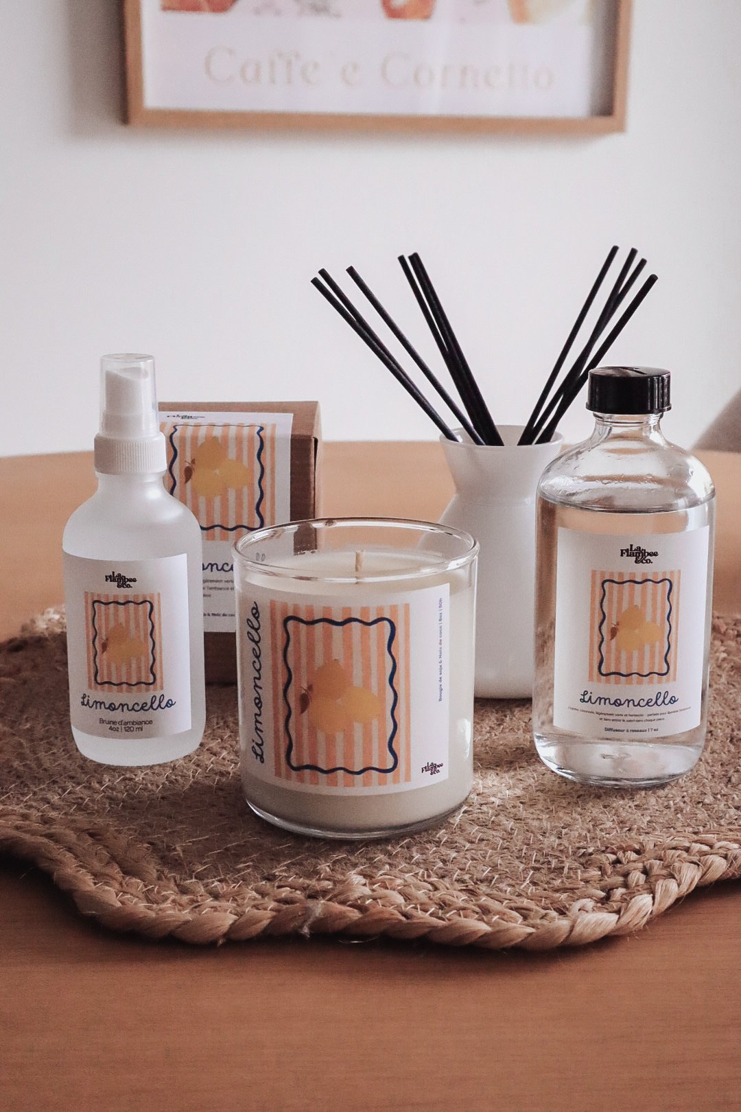 Différents produits de parfum ou d'ambiance appelés 'Limoncello' avec une bougie, un spray, une bouteille d'eau et un vase avec des bâtonnets, sur un tapis en rotin. Un tableau est en arrière-plan.