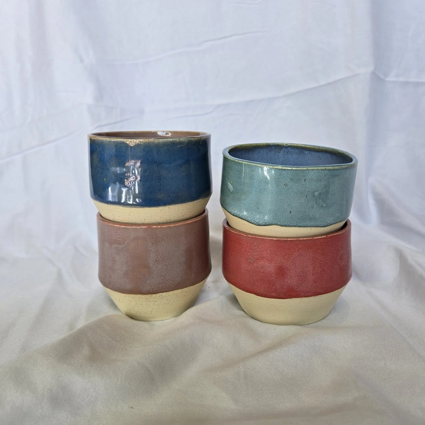 Six petits pots en céramique, empilés deux par deux, avec des couleurs pastel : bleu, vert et rose, posés sur un tissu blanc