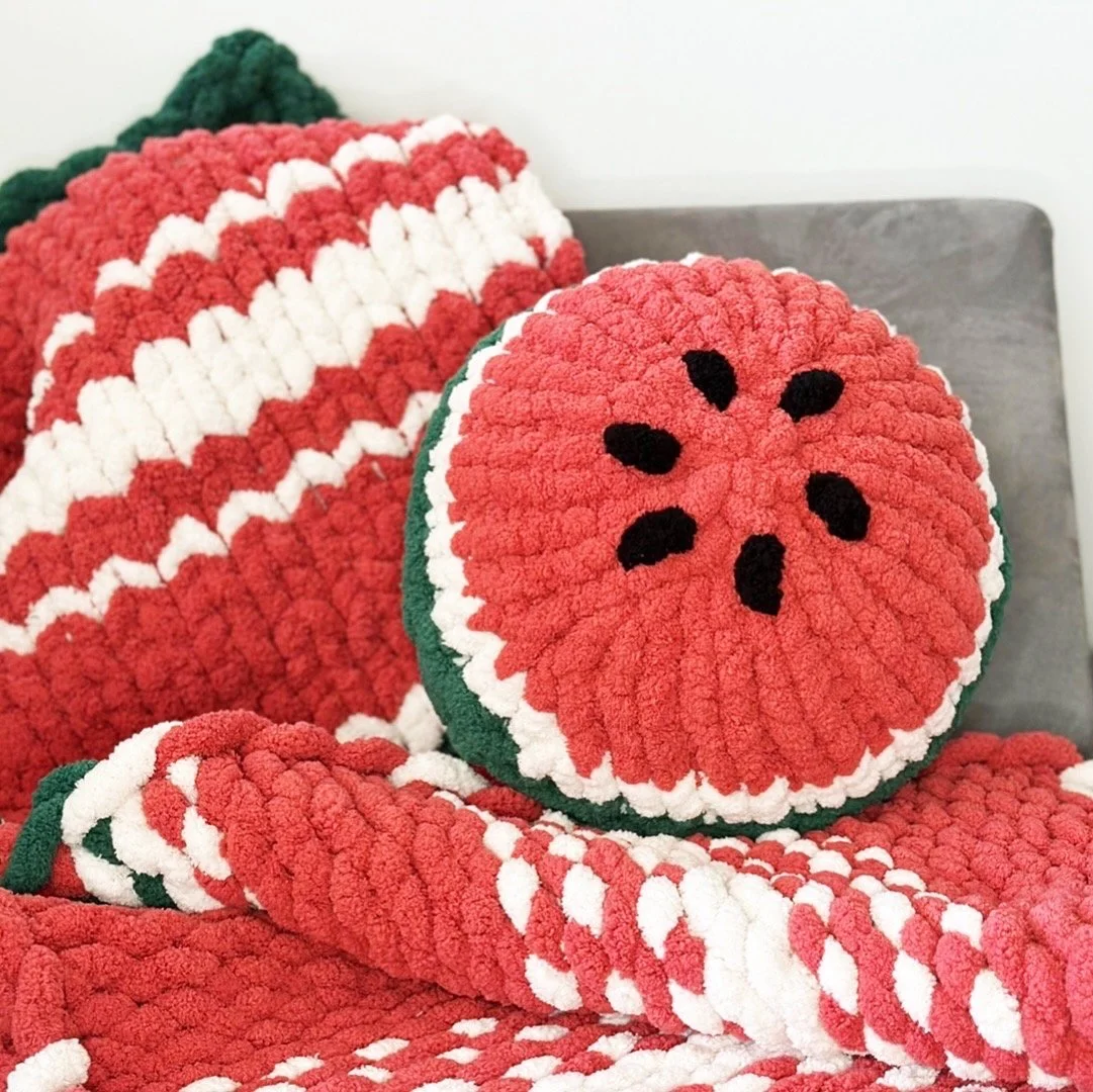 Coussin en forme de pastèque, avec des motifs rouges et noirs, posé sur une couverture en crochet rouge et blanc.