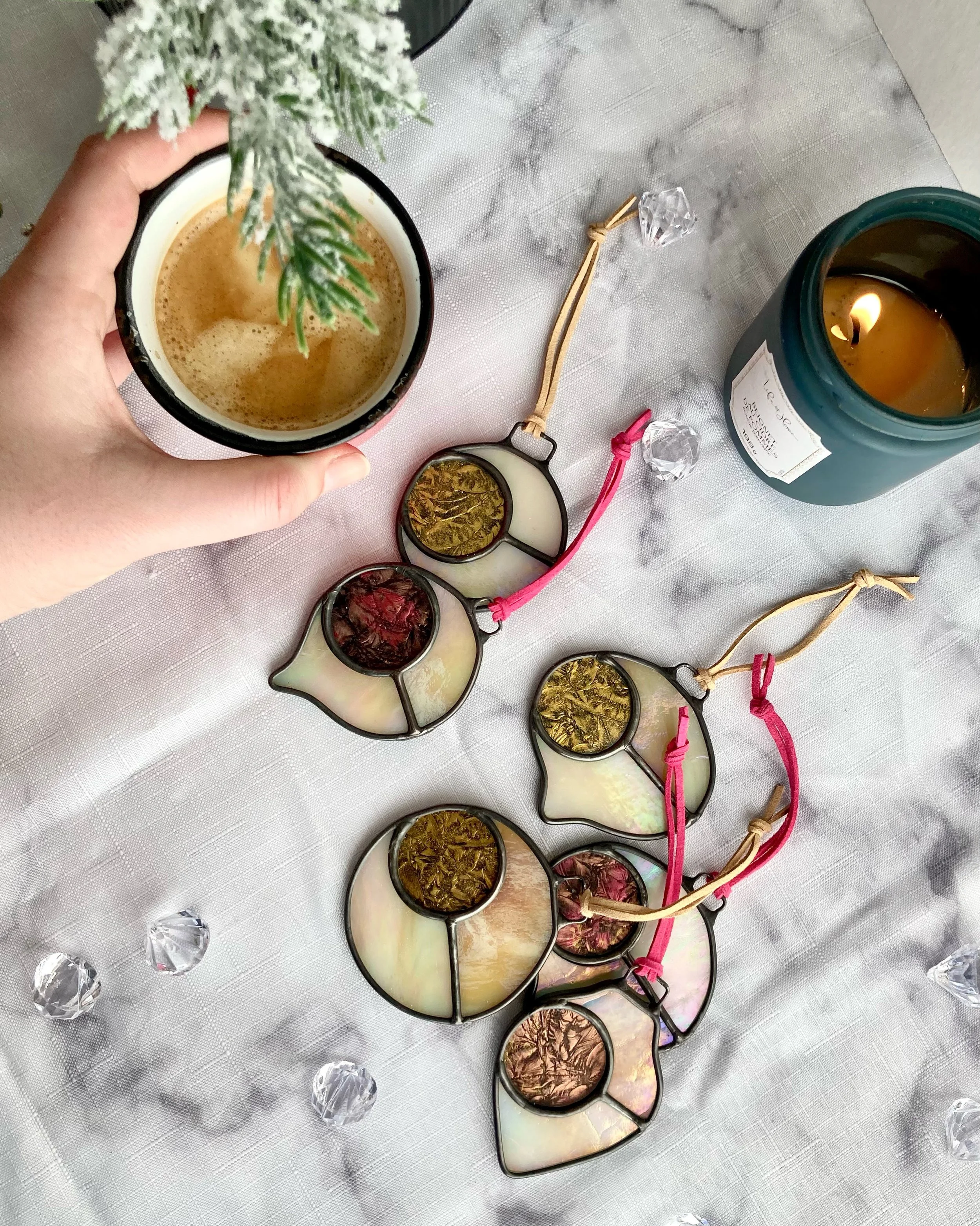 Une tasse de café ou de thé, un bouquet de fleurs faux saupoudrées de neige artificielle, une bougie allumée dans un pot en verre, et une collection de lunettes de soleil de différentes couleurs et formes, sur une table recouverte d'une nappe blanche en tissu.