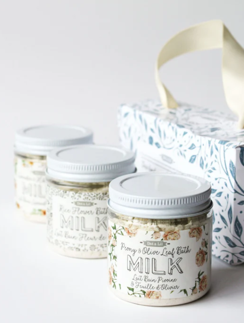 Trois petits pots de bain au lait avec des étiquettes illustrées, accompagnés d'un boîte cadeau en papier avec une poignée en ruban.