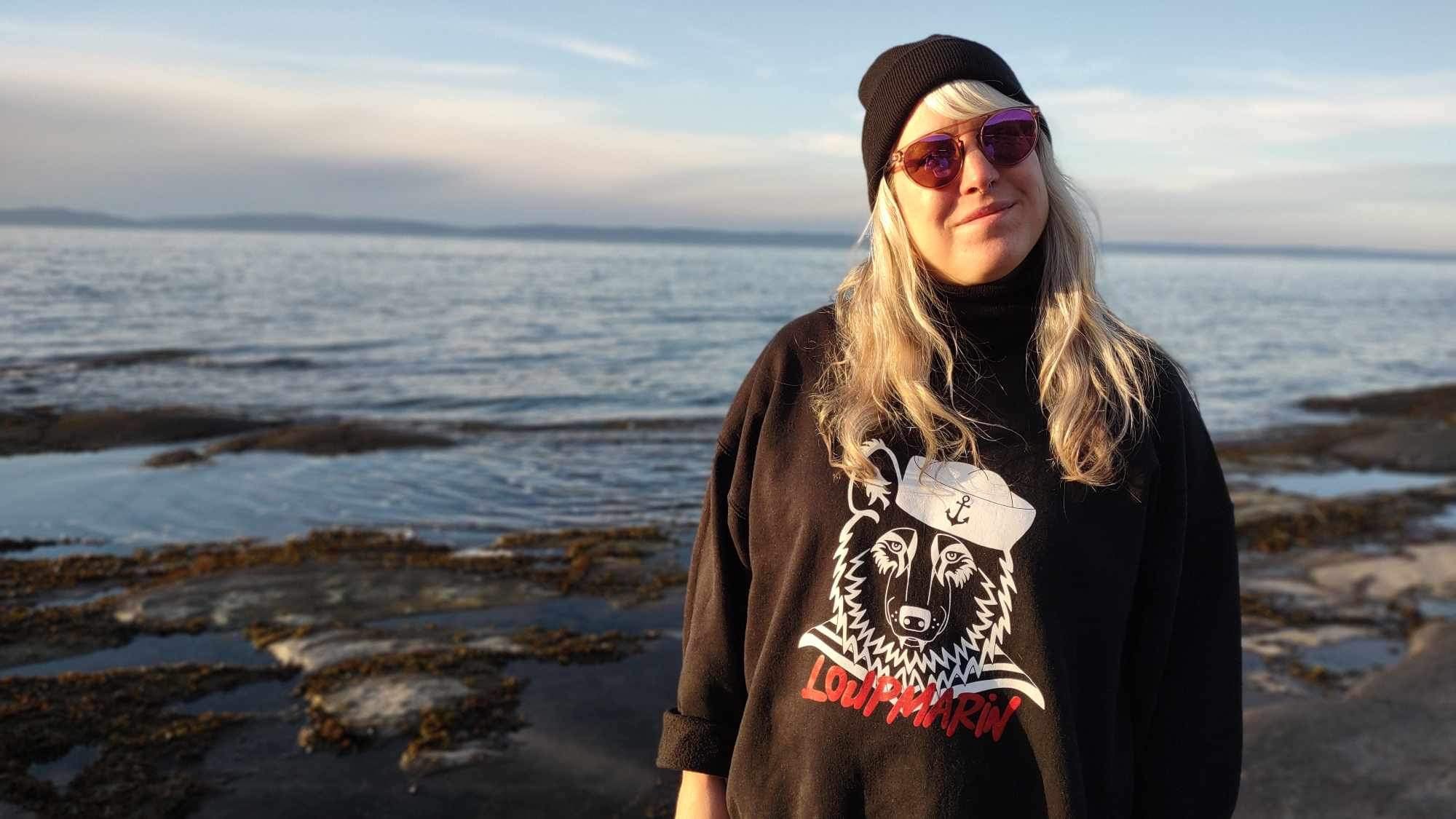 Jeune femme portant un bonnet noir, lunettes roses et un sweat noir avec un lion et le nom "Loupmarvin", debout près de la mer avec un ciel nuageux en arrière-plan.