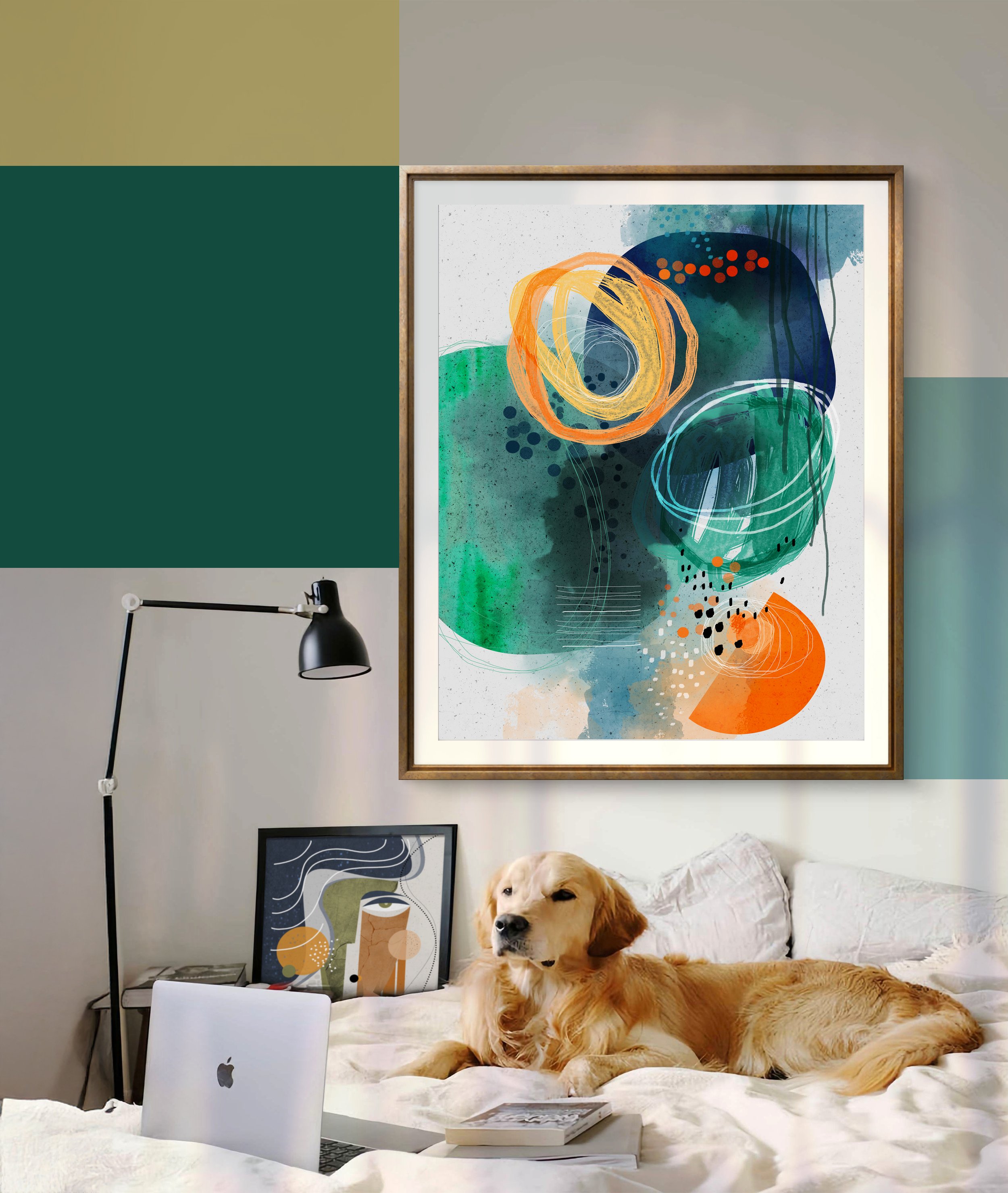 Un chien golden retriever allongé sur un lit blanc devant un ordinateur portable. Sur le mur, plusieurs œuvres d'art abstraites encadrées. À gauche, une lampe noire à bras articulé. Sur la table, une pile de livres et des papiers.