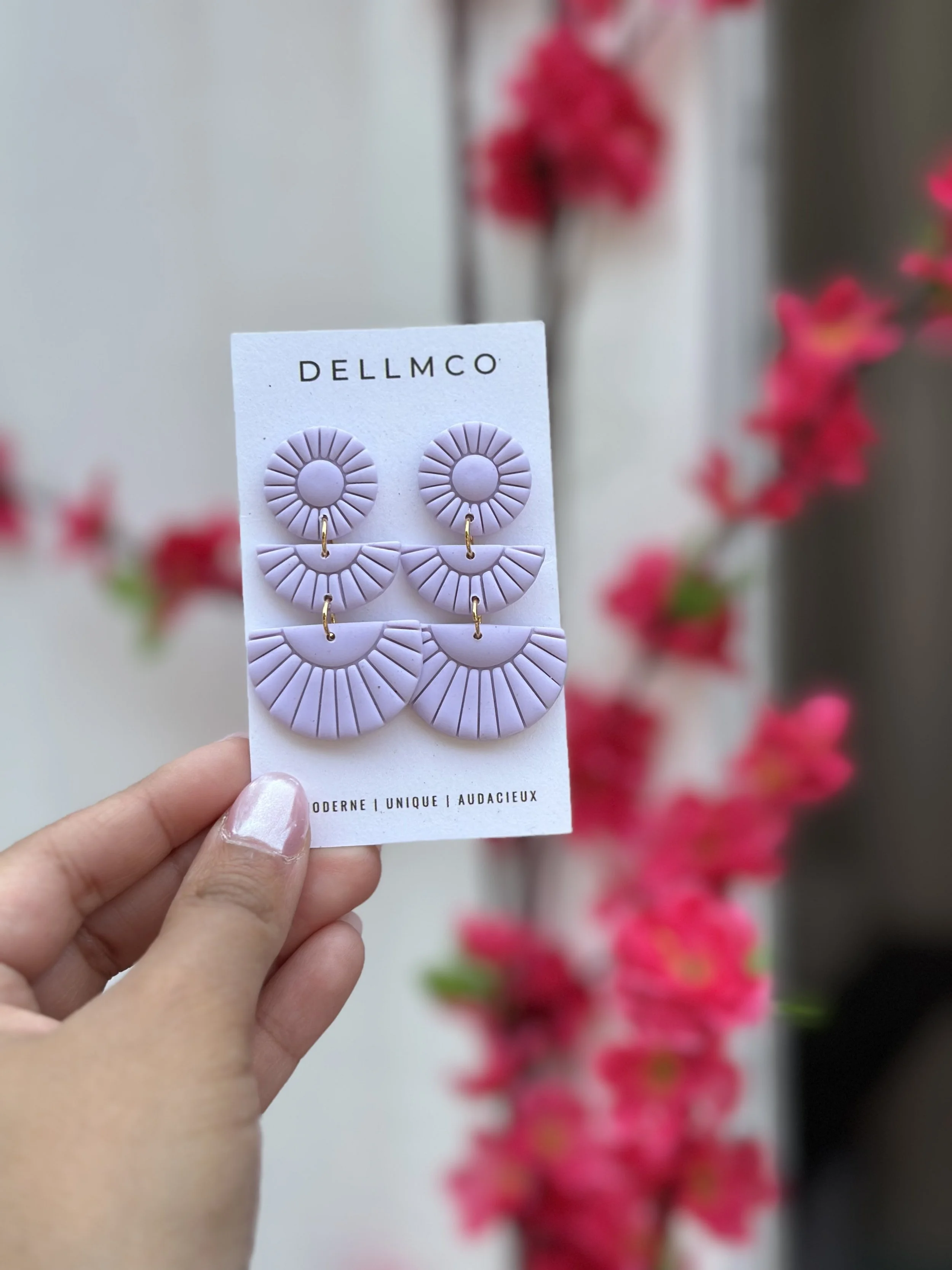 Boucles d'oreilles violettes en forme de cercles avec des rayures, présentées sur un support blanc avec la marque 'DELLMCO', contre un fond flou de fleurs roses.