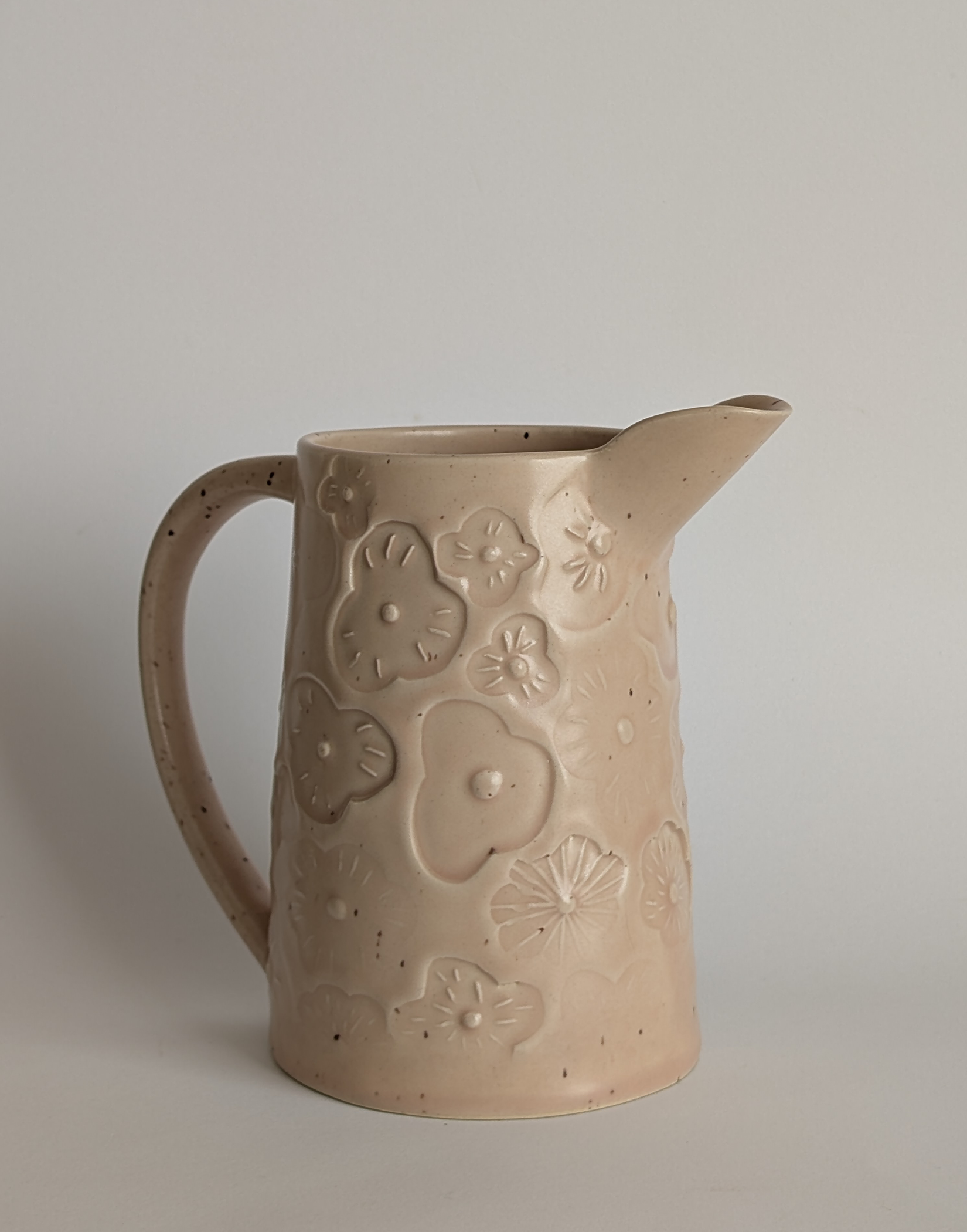 Une cruche en céramique beige avec un motif de fleurs en relief, placée contre un fond blanc.
