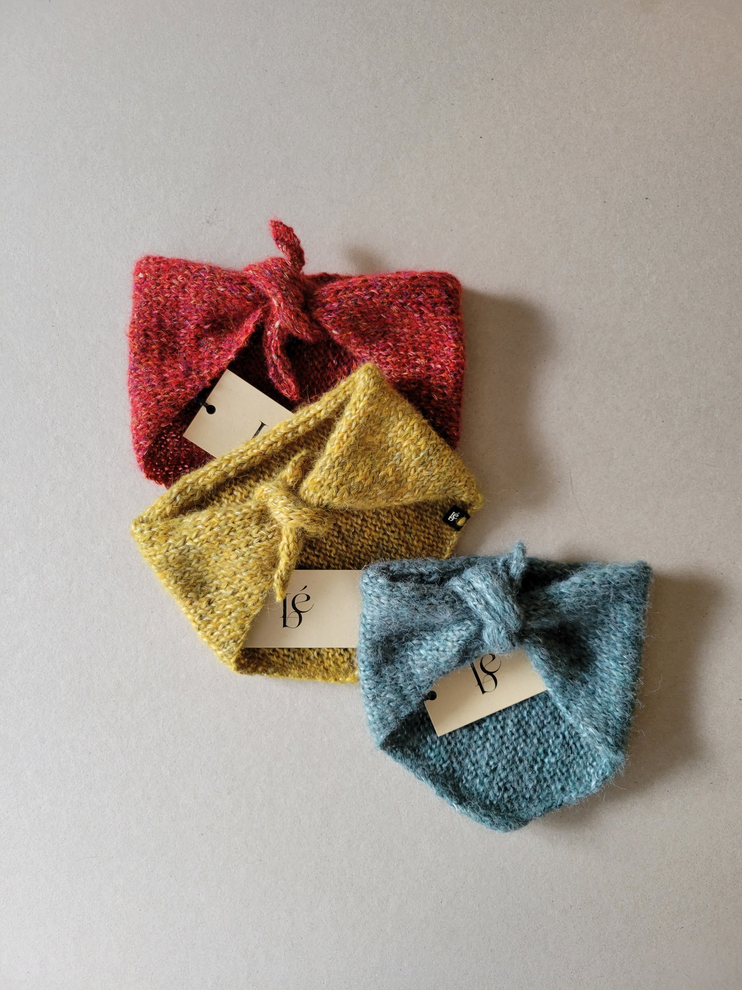 Trois petits bandeaux ou serre-heads en laine de différentes couleurs, rouge, jaune et bleu, chacun avec un tag blanc portant une lettre ou un logo.