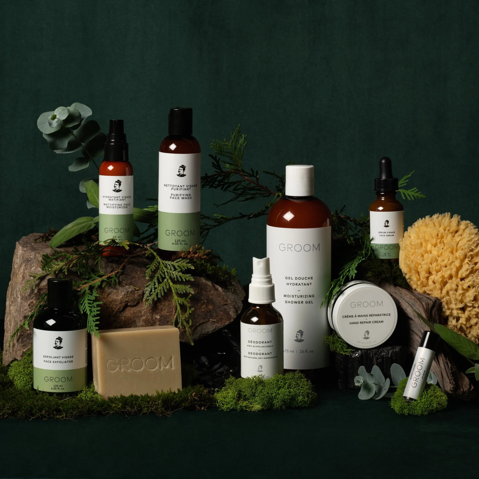 Gamme de produits de soins pour homme GROOM, comprenant des bouteilles de sérum, déodorant, gel douche, crème, savon, spray, présentée sur un socle en bois avec des éléments naturels tels que des branches de conifère, des feuilles vertes, et une éponge naturelle, avec un fond vert foncé.