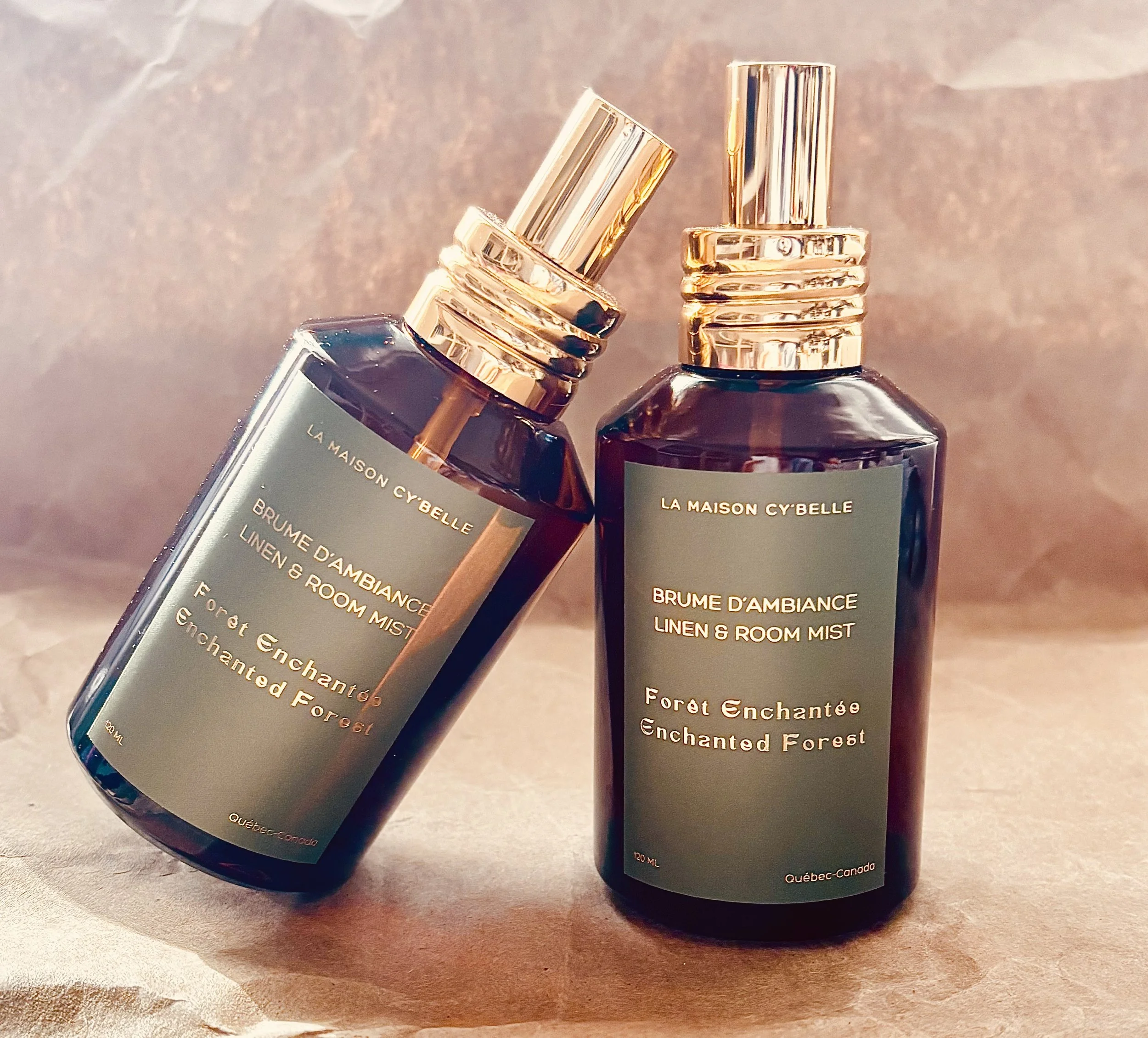 Deux bouteilles de parfum de couleur ambre avec des bouchons en or rose, étiquetées « Forêt Enchantée » et « Brume d'Ambiance Linen & Room Mist », posées sur une surface en bois.