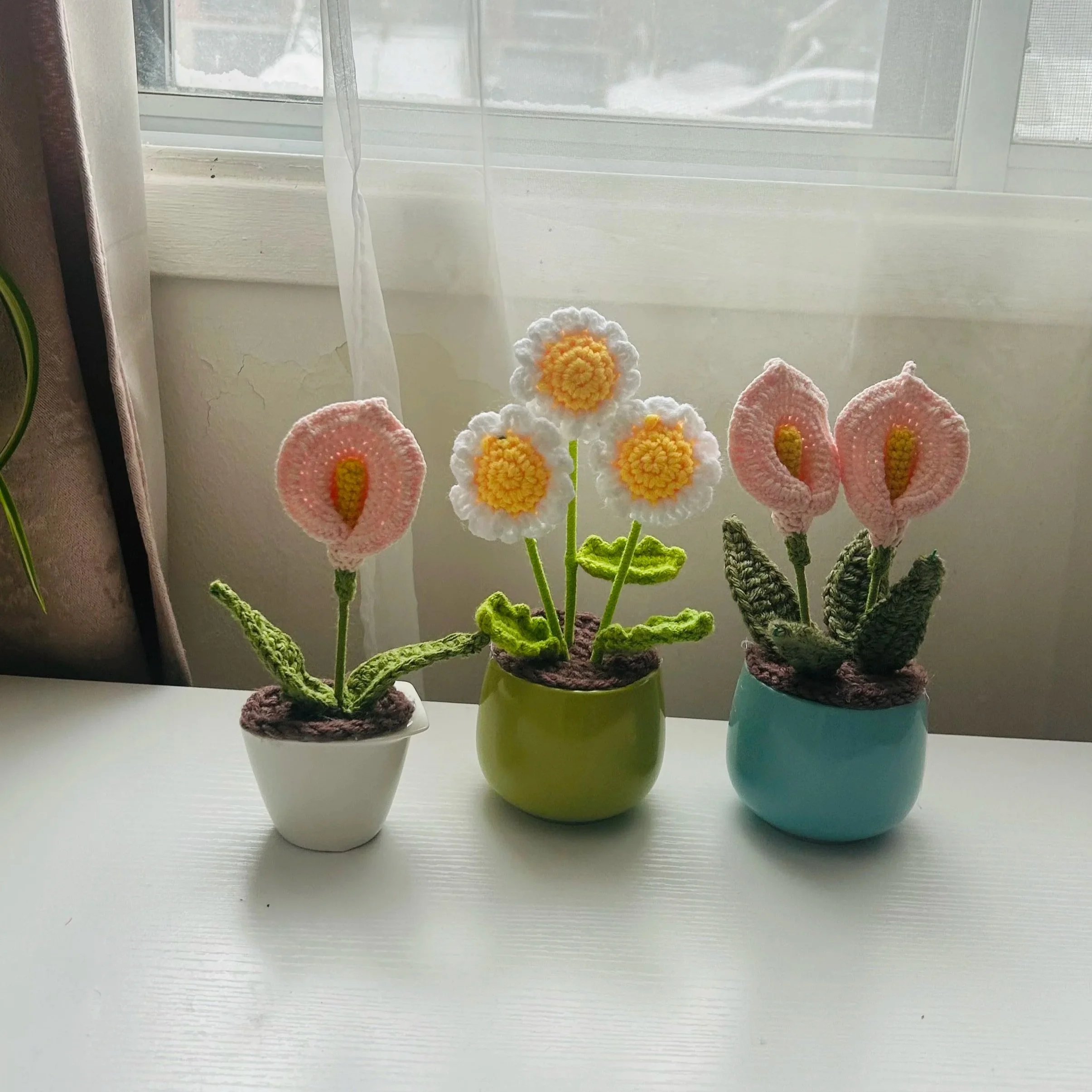 Vases avec des fleurs en crochet, placés sur une table blanche devant une fenêtre avec des rideaux transparent, un mur blanc avec des fissures.