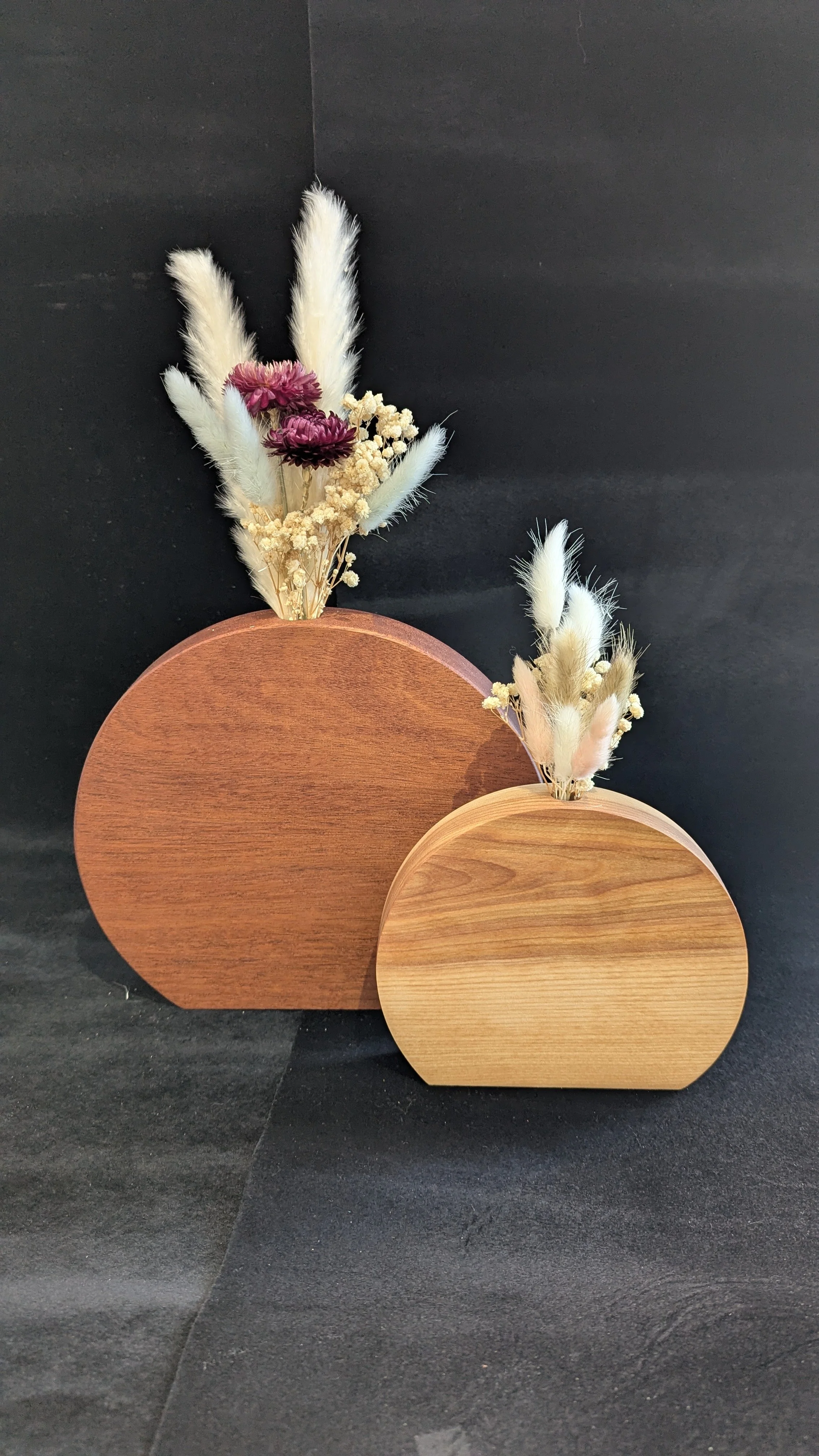 Deux décorations florales avec des vases en bois circulaires, contenant des fleurs séchées blanches, roses et violettes, contre un fond noir.