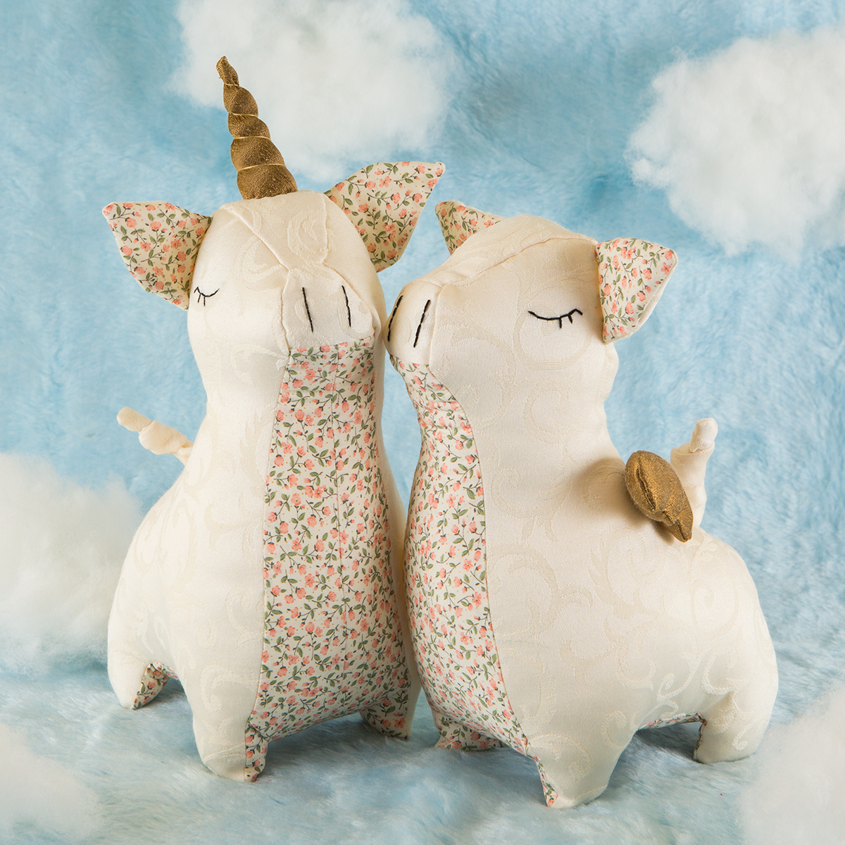 Deux peluches de licornes avec un corps en tissu crème, des oreilles en tissu floral, des cornes en tissu doré, et des ailes en tissu floral, sur un fond nuageux bleu.