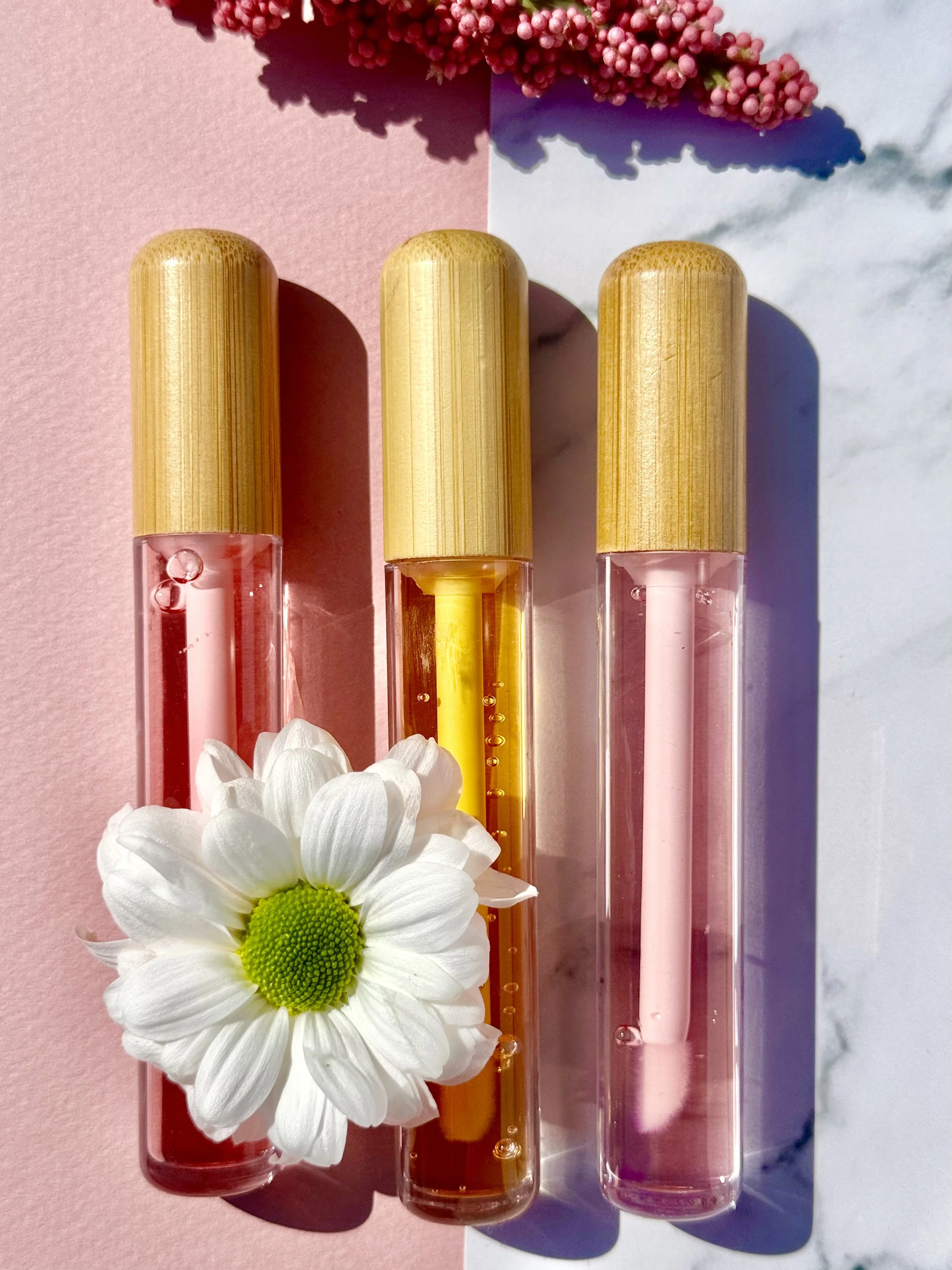 Trois flacons de liquide coloré (rose, orange et rose pâle) alignés, avec une fleur blanche à gauche et un bouquet rose en arrière-plan, sur une surface combinée rose et blanche.
