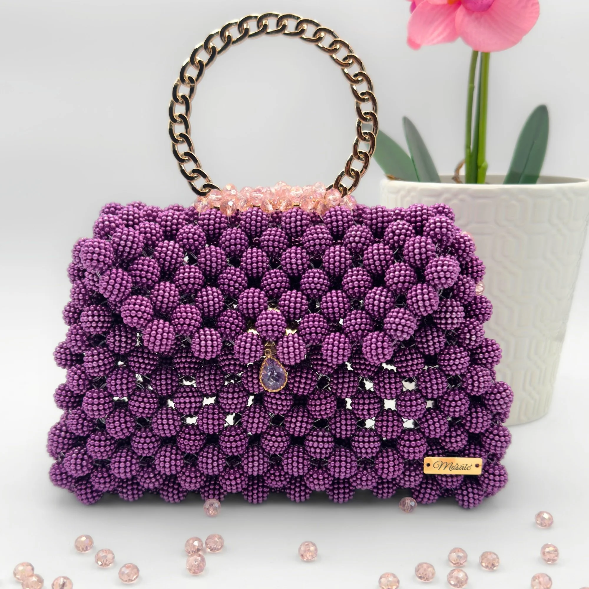 Sac à main violet en perles de rocaille avec une chaîne dorée et des cristaux roses. À côté, pot blanc avec une orchidée rose. Perles roses dispersées autour.
