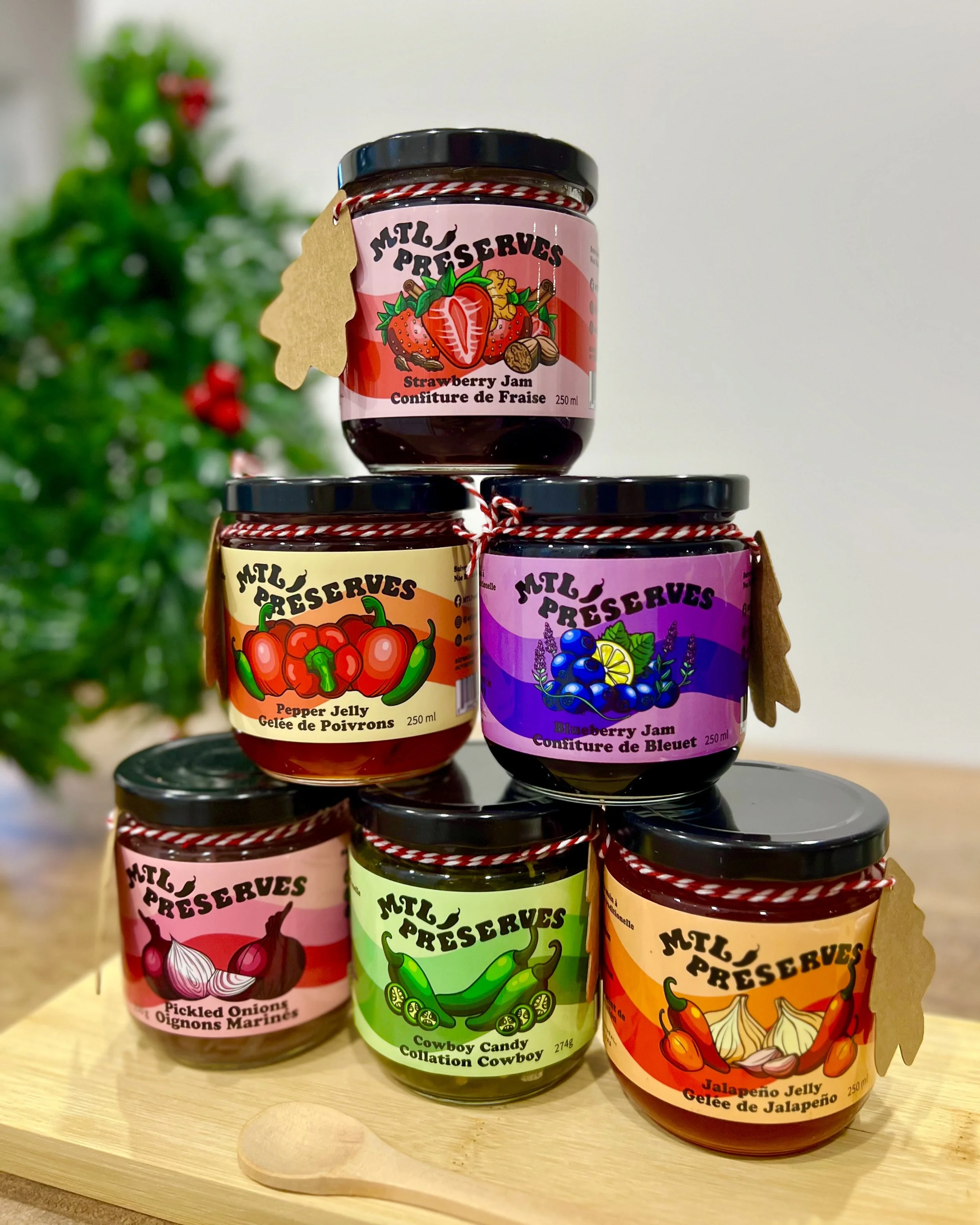 Décorations de Noël avec plusieurs pots de confitures de fruits, dont gelée de fraise, gelée de poivrons, confiture de bleuet, gelée de jalapeño, et collation cowboy, placés sur une table en bois.