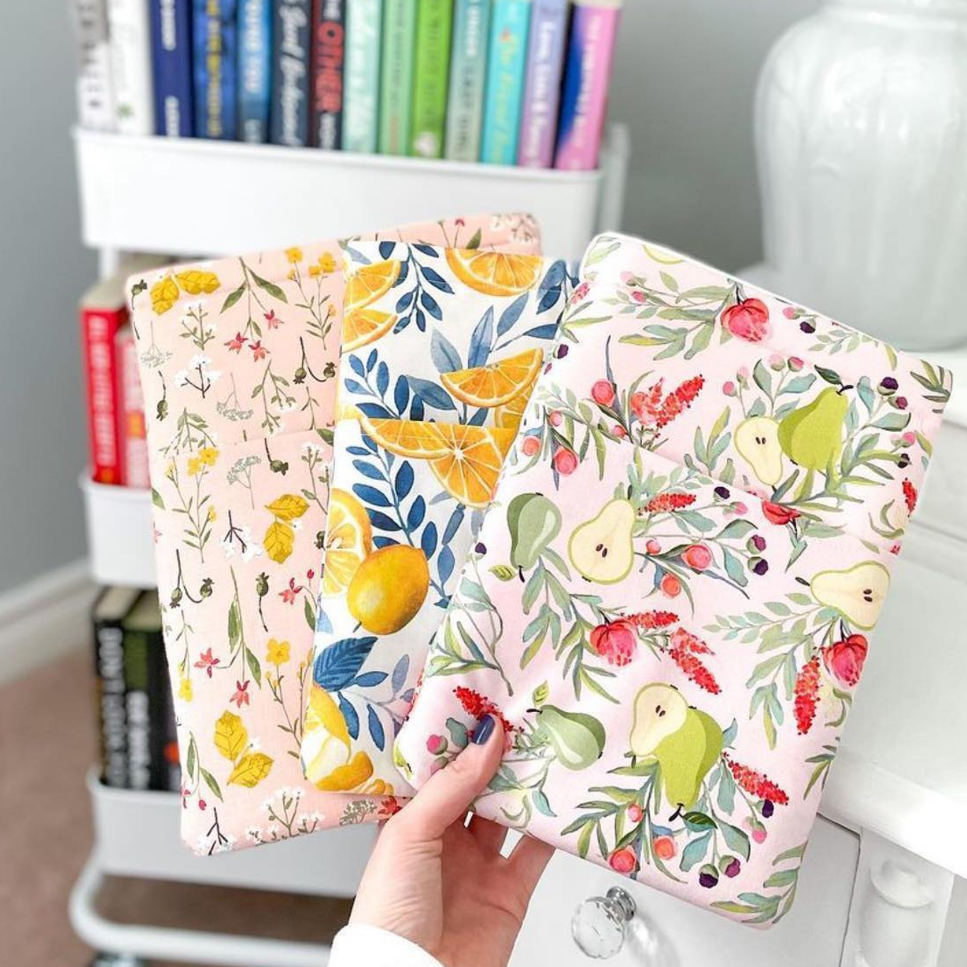 Main dans une main tenant trois papiers ou cartons aux motifs floraux et fruités, avec en arrière-plan une étagère blanche contenant des livres colorés, et une grosse jarre blanche sur une table ou un meuble blanc.