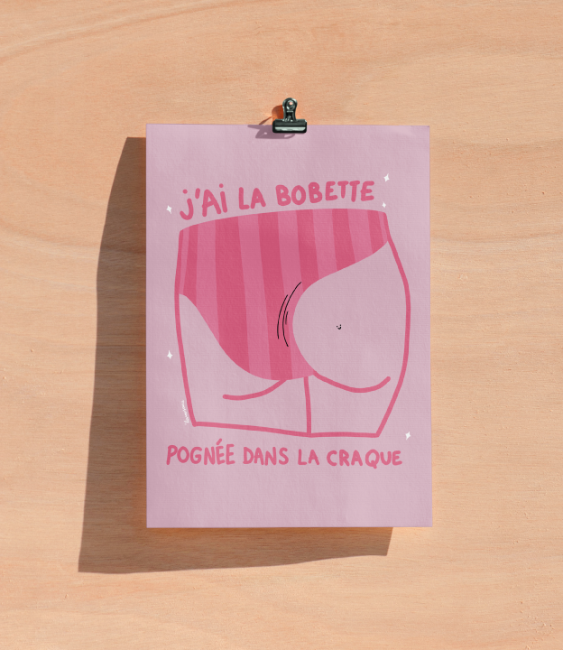 Une illustration humoristique d'une fesse dans un style dessin animé avec le texte "J'ai la bobette pognée dans la craque" sur une feuille rose attachée par une pince sur un fond en bois clair.