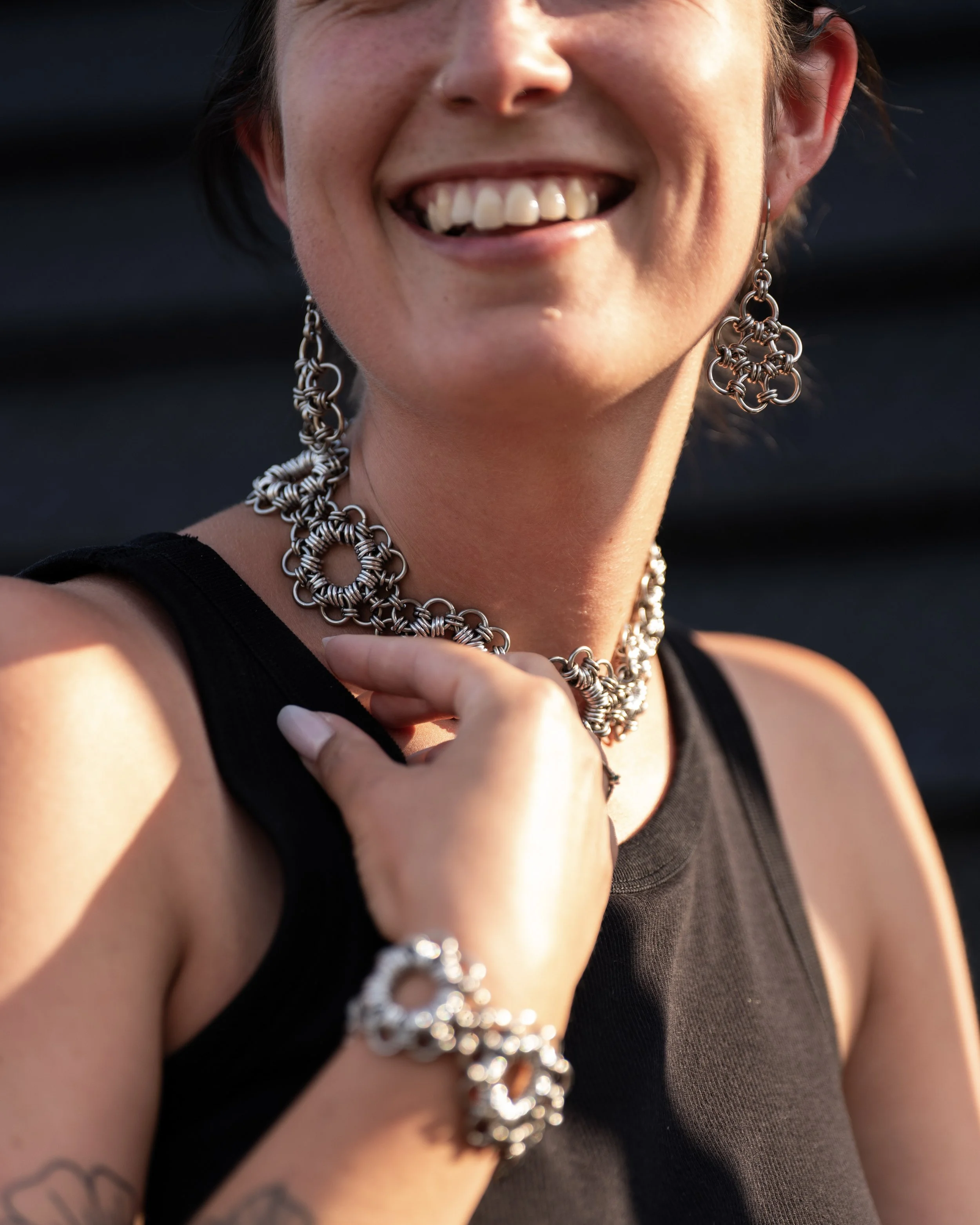 Une femme souriante portant des bijoux en métal, comprenant un collier, une paire de boucles d'oreilles et un bracelet, vêtue d'un haut noir.